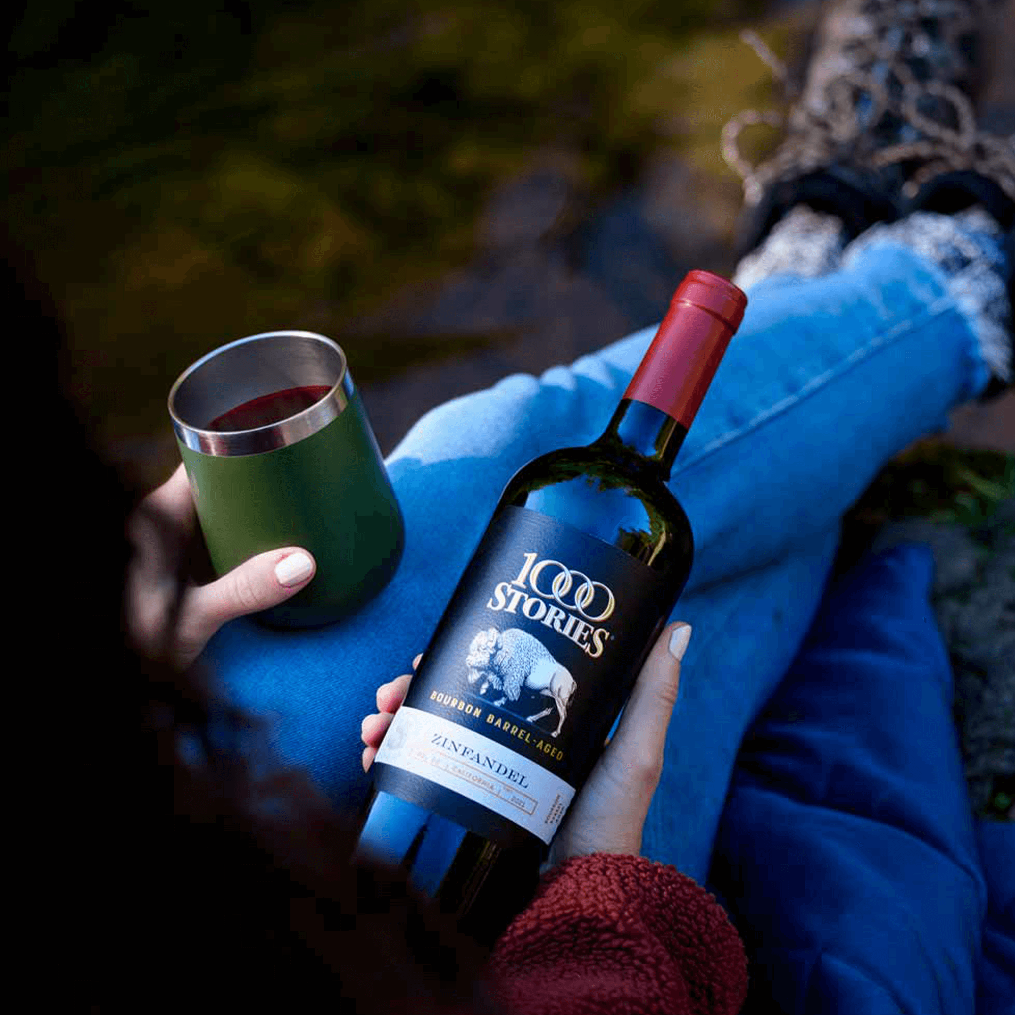 A person enjoying 1000 Stories Bourbon Barrel Aged Zinfandel while seated outdoors by a stream.