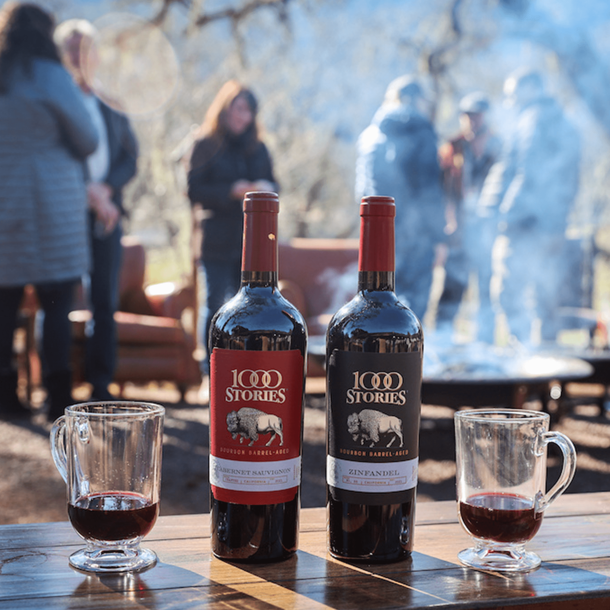 Bottles of 1000 Stories Bourbon Barrel Aged Zinfandel and Cabernet Sauvignon with glasses of wine outdoors.