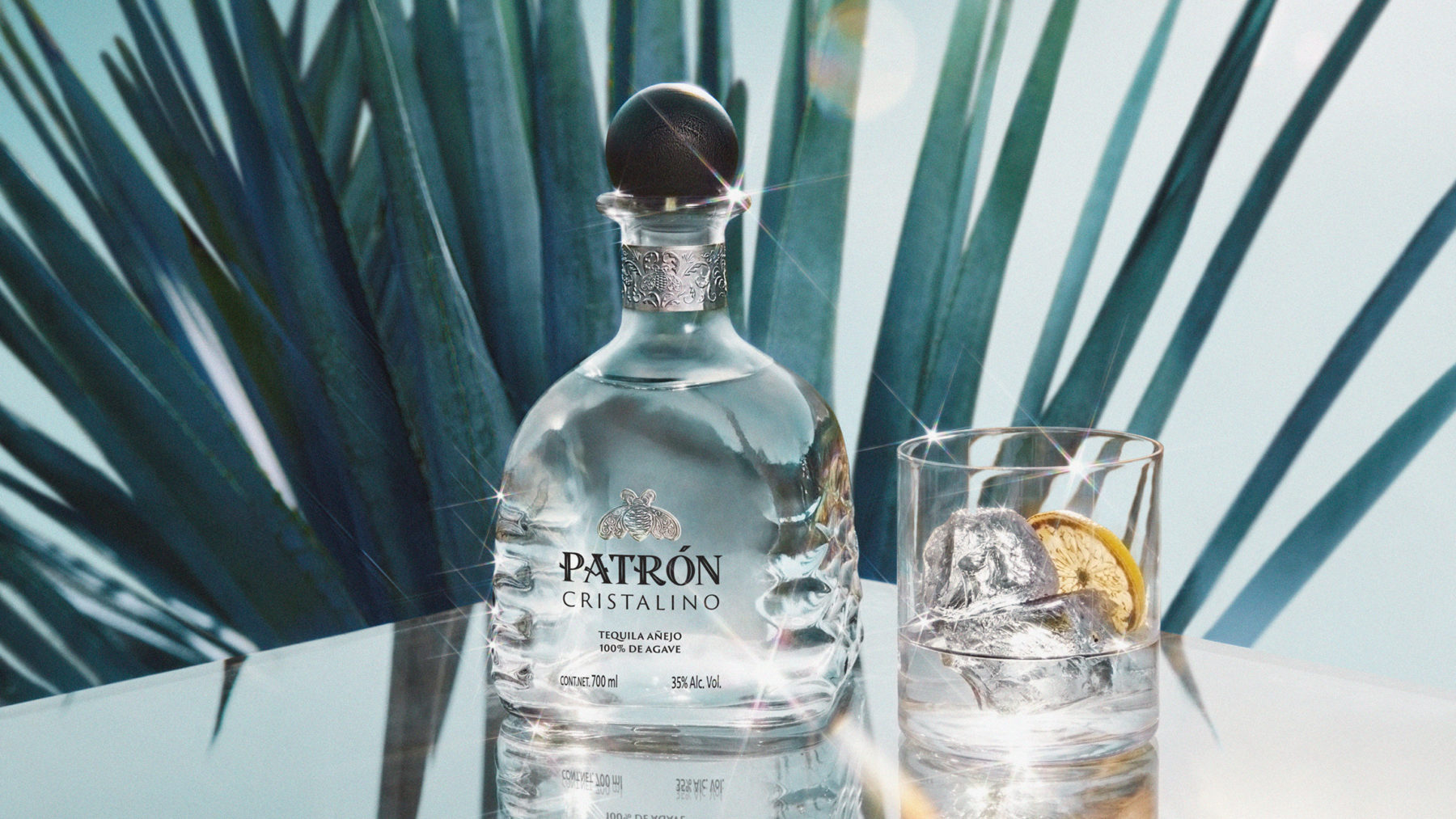 Patrón Cristalino Añejo Tequila bottle and glass with ice, garnished with lemon, set against a vibrant plant background.