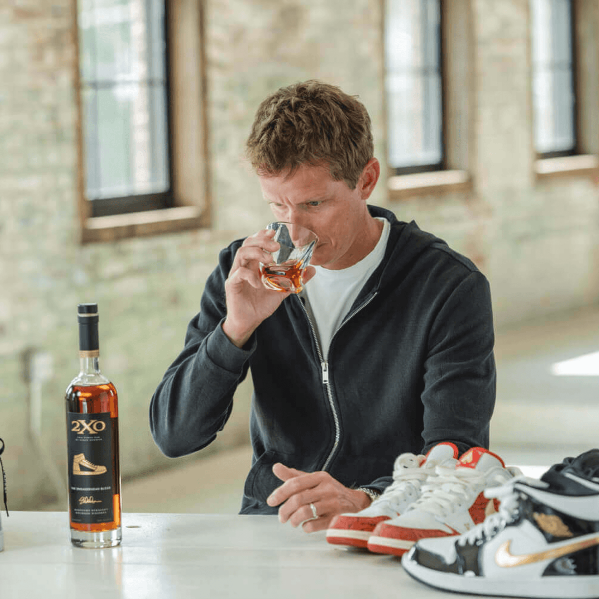 Man enjoying 2XO The Sneakerhead Blend bourbon next to stylish sneakers, showcasing sneaker culture and collectible fashion.