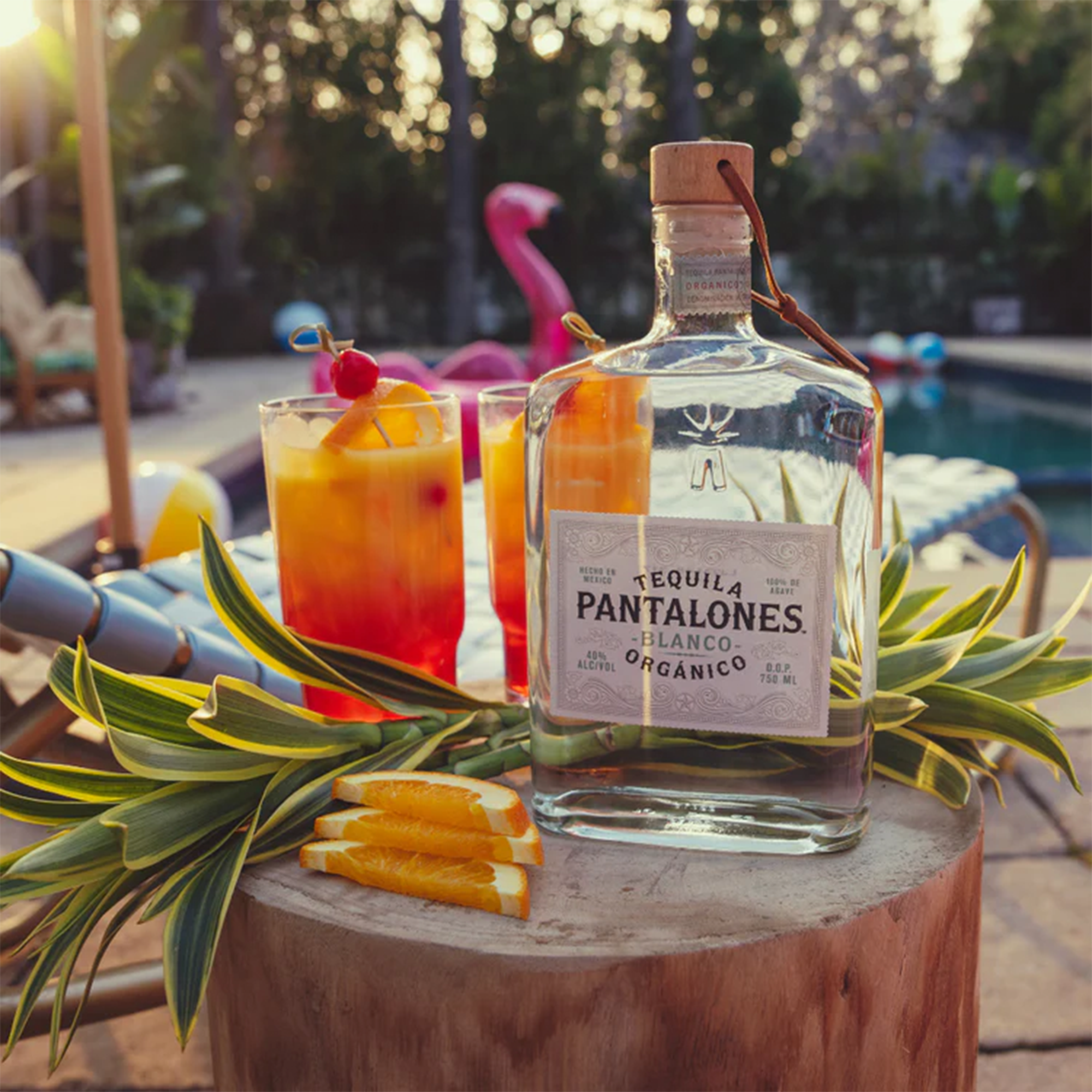 Pantalones Blanco Tequila bottle with tropical cocktails and fresh oranges by a poolside, embodying summer vibes.