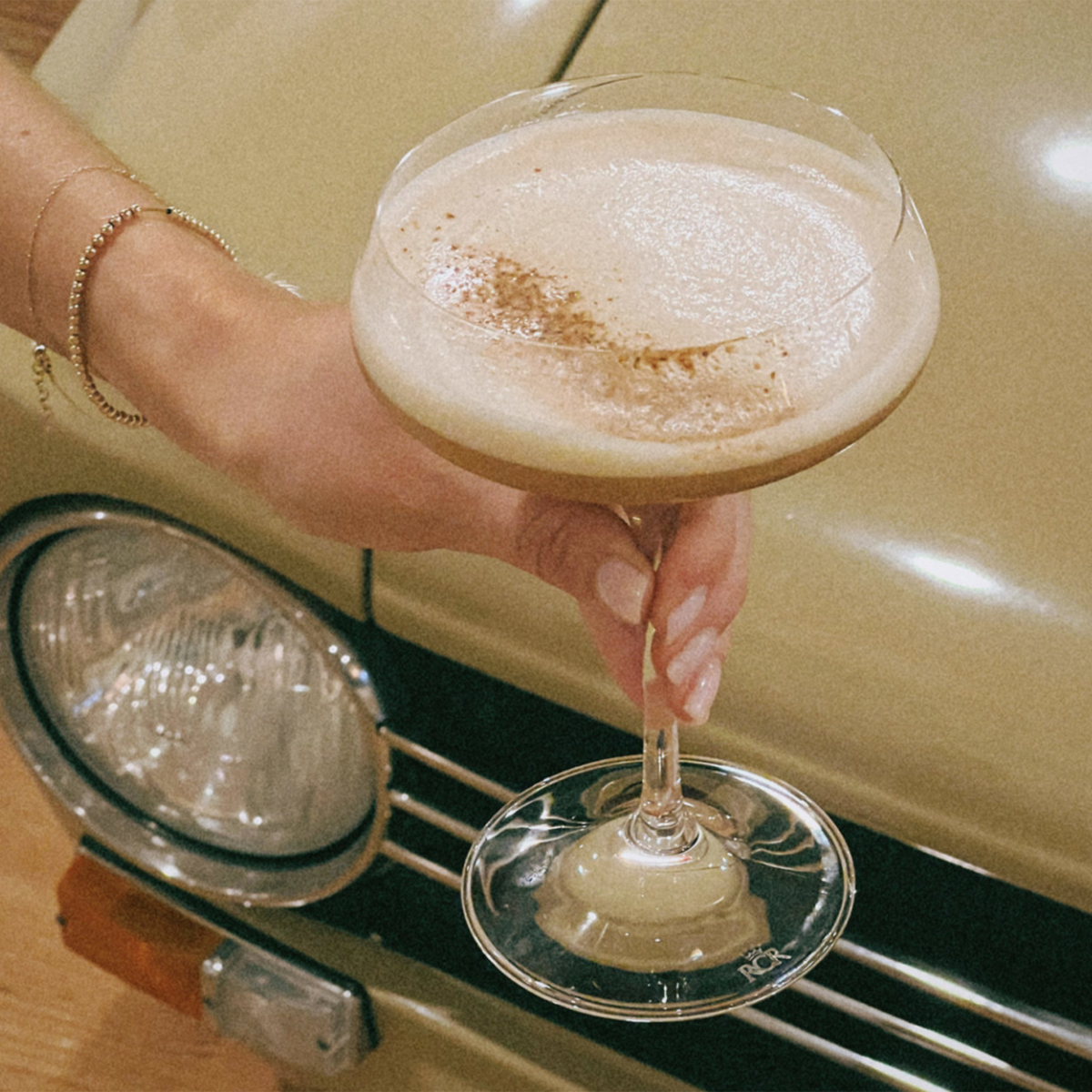 A hand holding a cocktail glass with a frothy drink, set against a vintage car background.