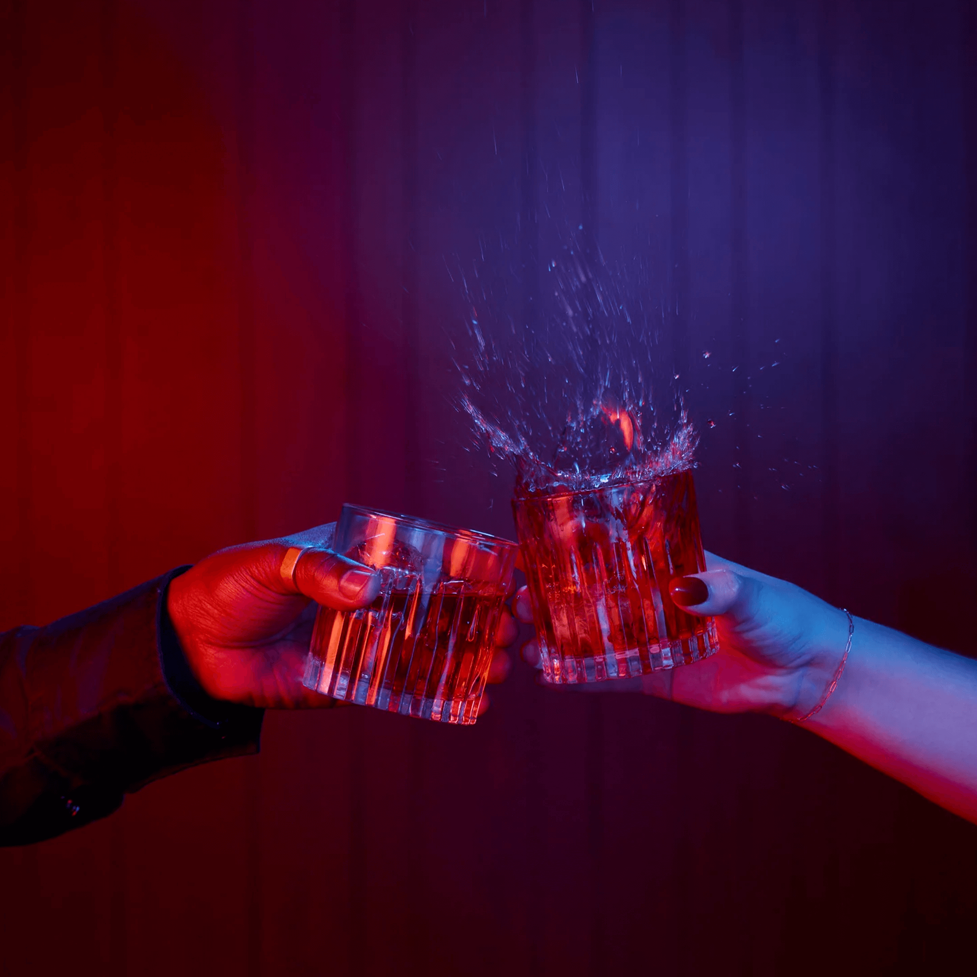 Two hands clinking whiskey glasses, capturing a splash against a vibrant red and blue backdrop.