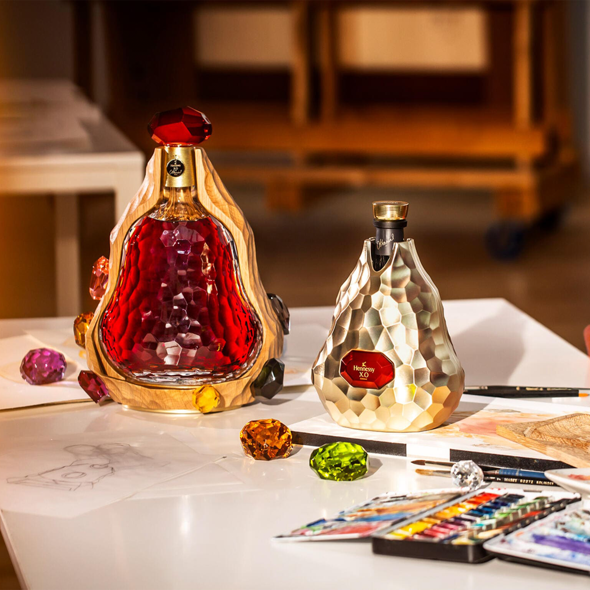 Hennessy x Jean-Michel Othoniel X.O. Cognac limited edition bottles on a table with colorful gems and art supplies.