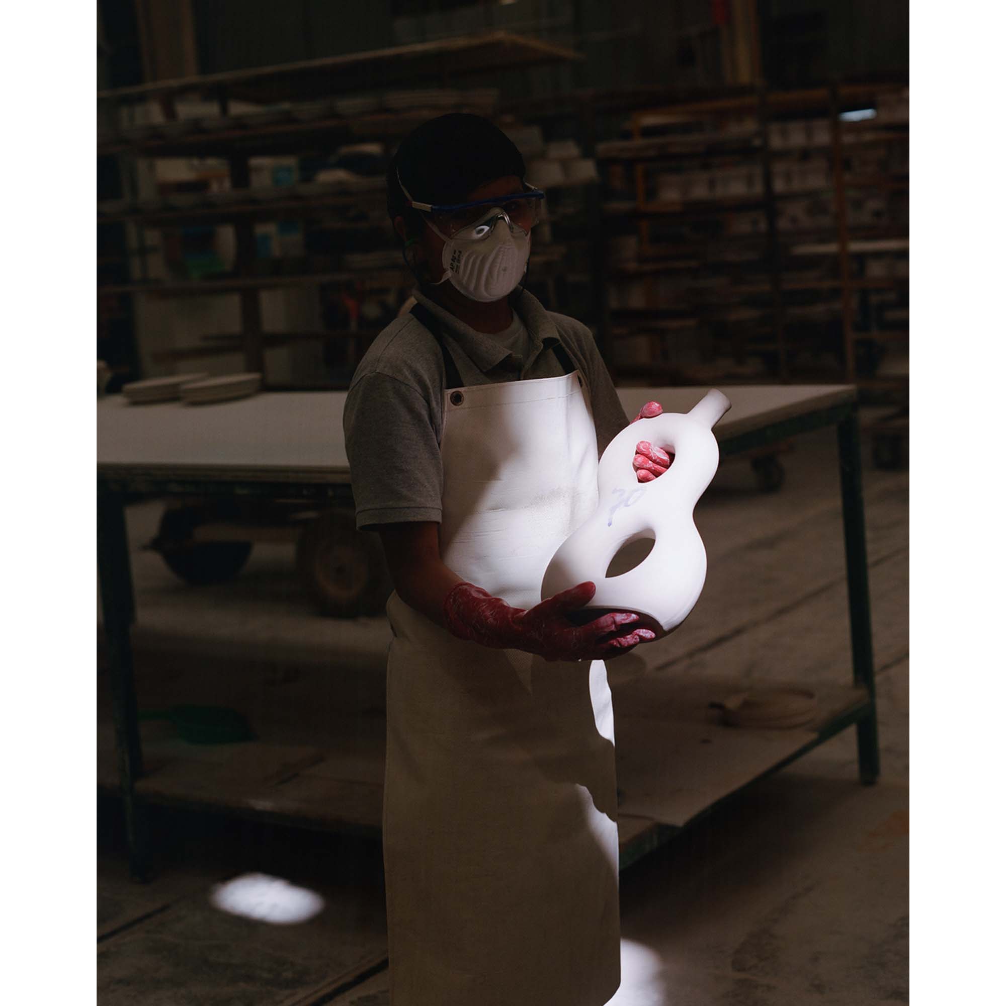 Craftsperson holding a white guitar mold in a workshop, wearing a mask and apron.