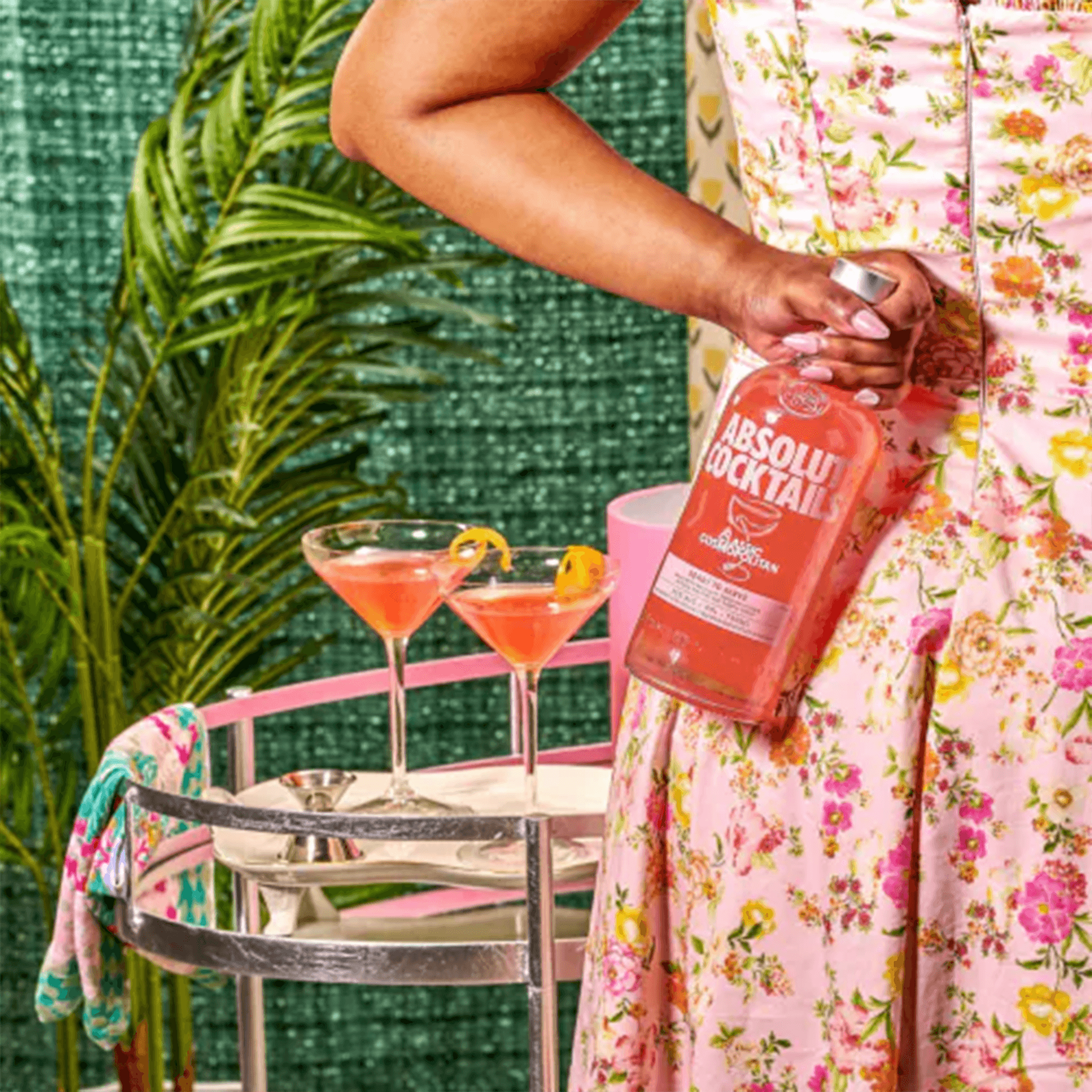 Woman in a pink floral dress holding Absolut Classic Cosmopolitan cocktail bottle beside two served drinks.