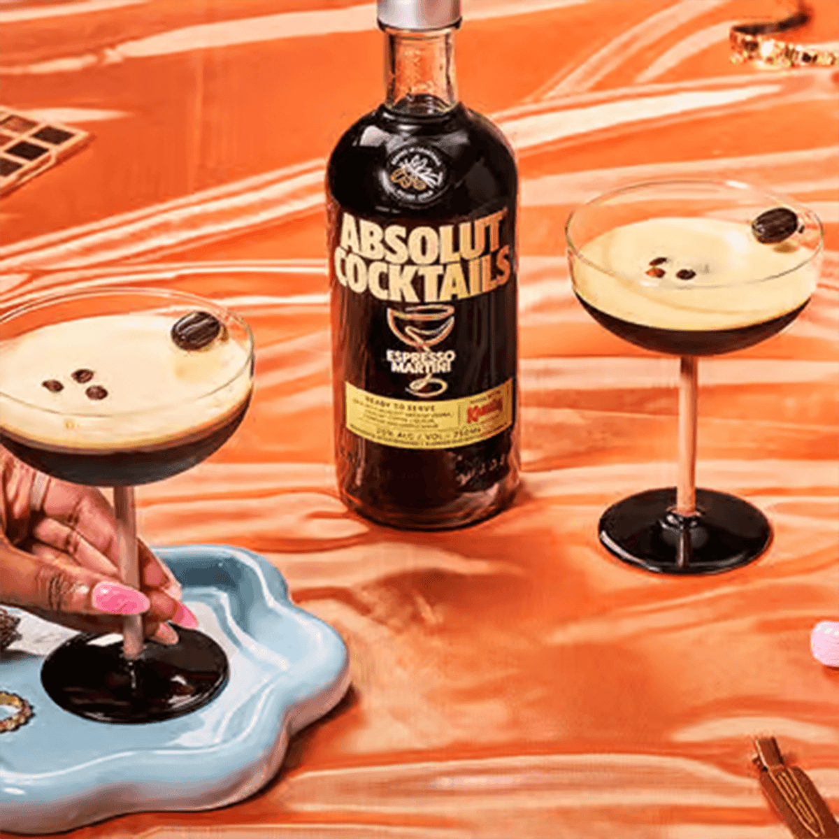 Absolut Cocktails Espresso Martini bottles with two glasses on an orange table backdrop, featuring coffee beans decor.
