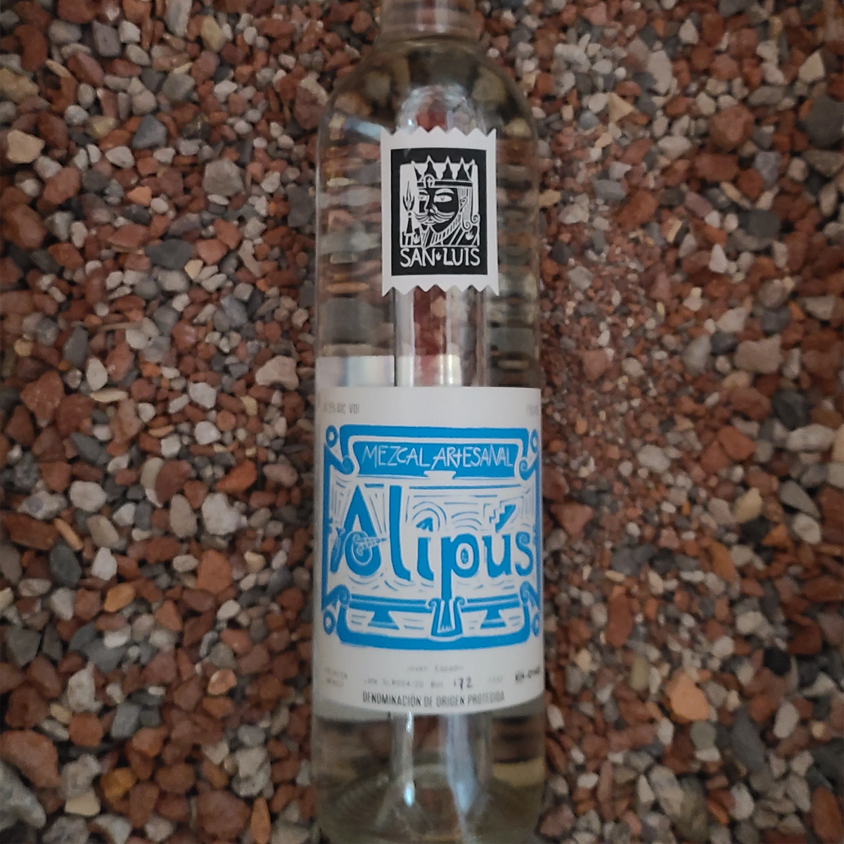 Alipus San Luis Del Rio Mezcal bottle on gravel, showcasing artisanal craftsmanship and vibrant design.