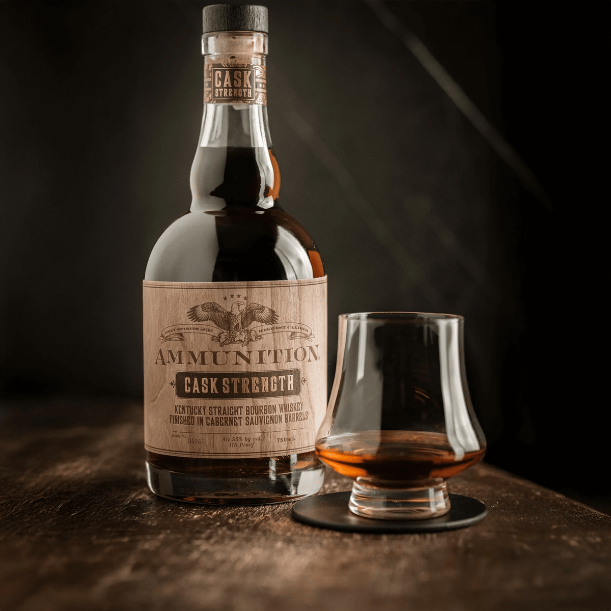 Ammunition Bourbon Cask Strength bottle with a glass showcasing rich amber whiskey on a wooden surface.