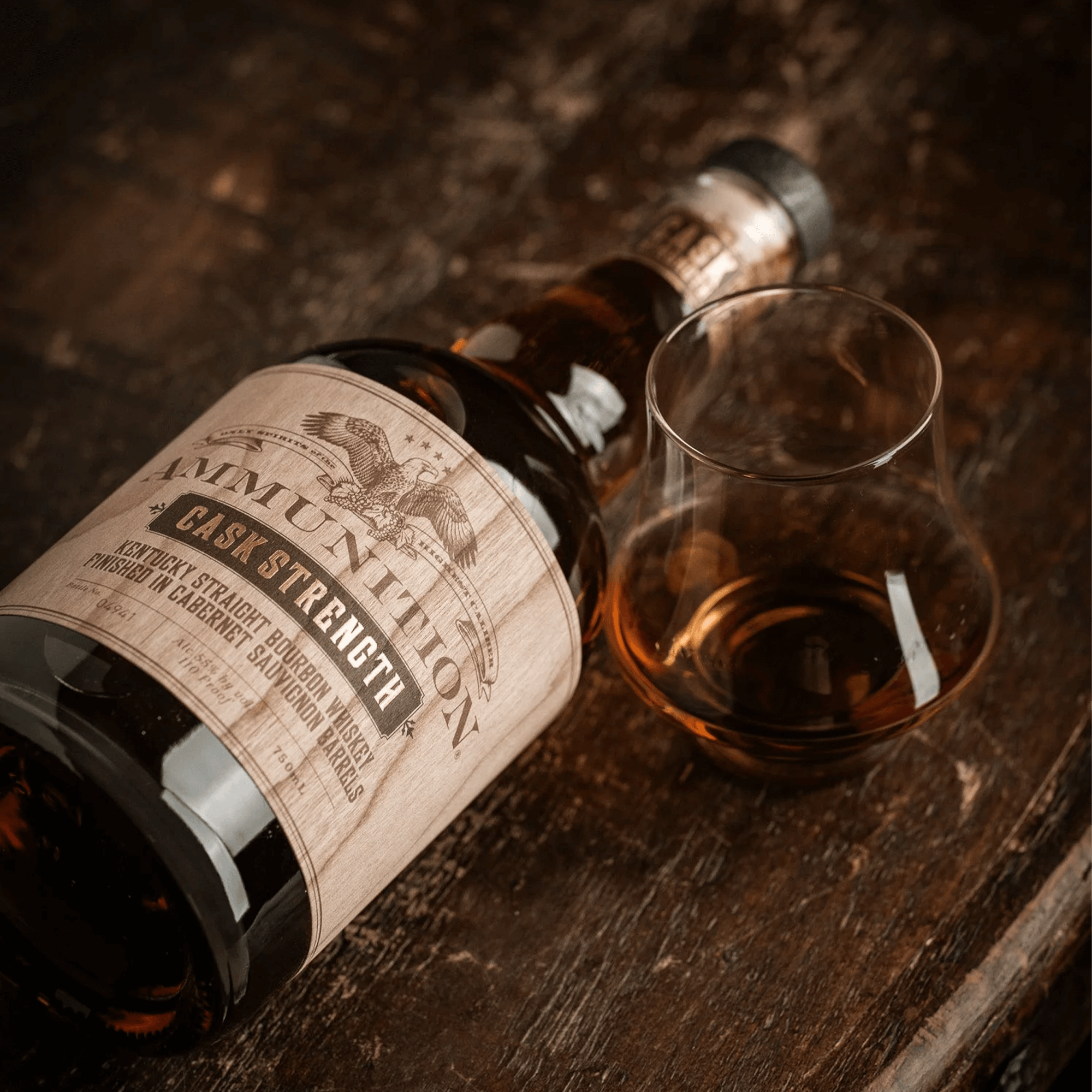 Ammunition Bourbon Cask Strength bottle beside a tasting glass filled with amber bourbon on a rustic wooden surface.