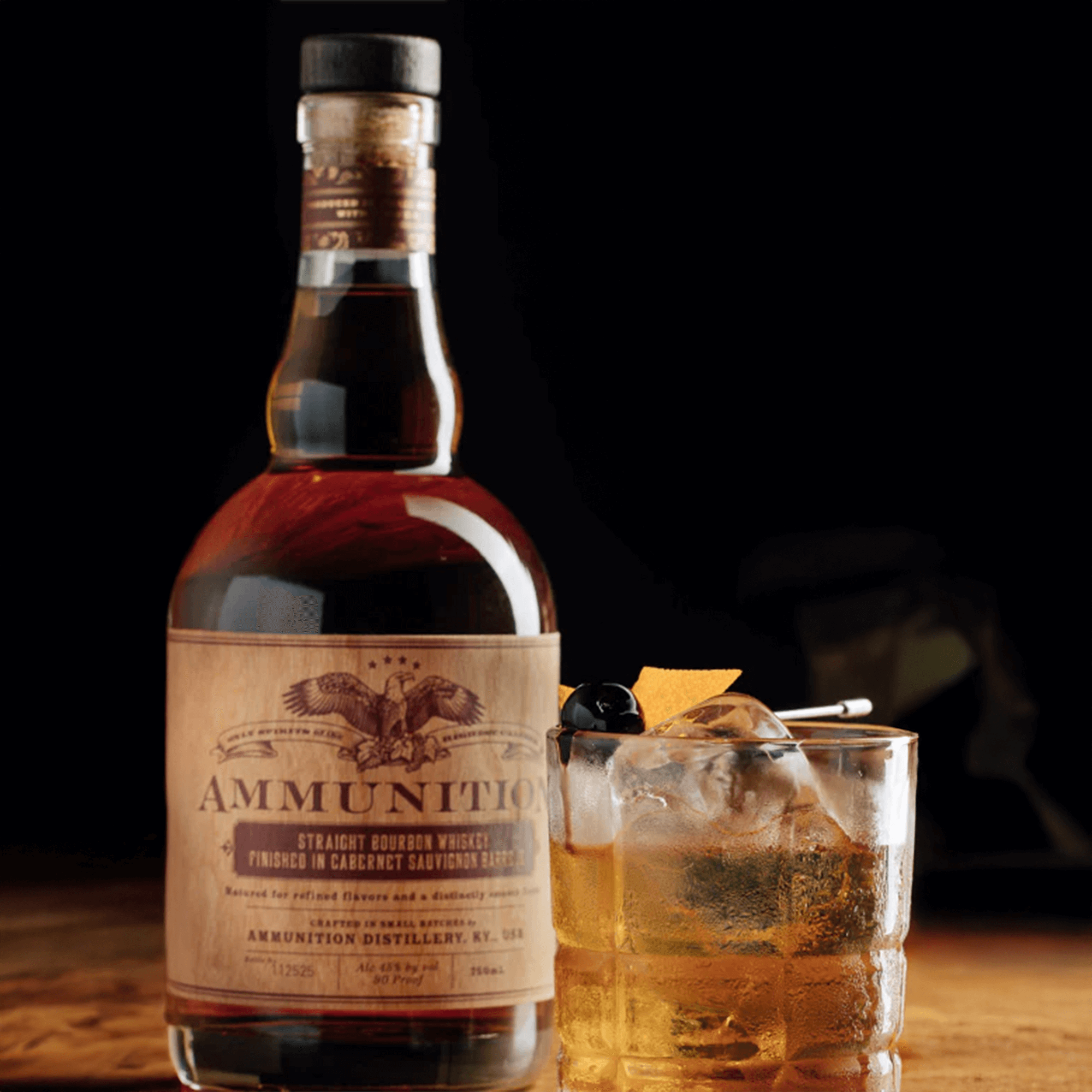 Ammunition Straight Bourbon Whiskey bottle with cocktail garnished with cherry and orange peel, showcasing its rich amber color.