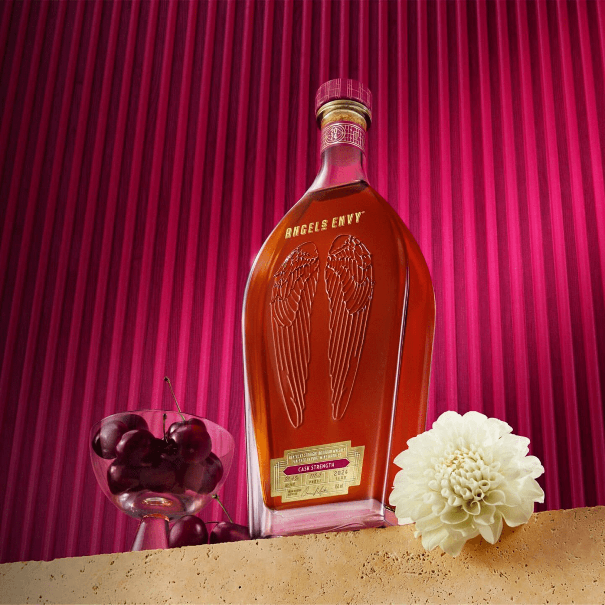 Angels Envy Cask Strength bourbon bottle with cherries and a flower, set against a vibrant pink striped background.