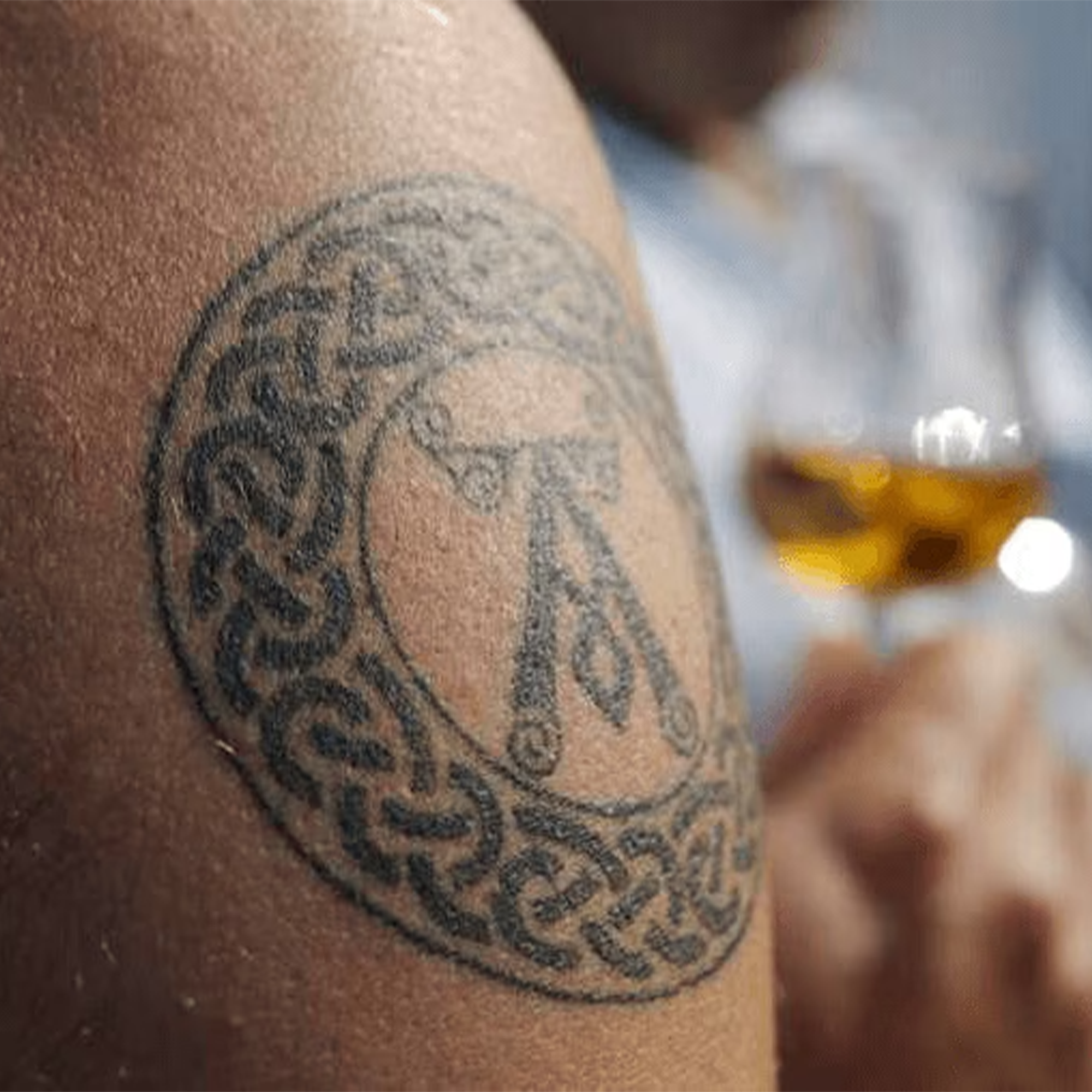 Close-up of a Celtic tattoo on an arm with a glass of whiskey in the background.