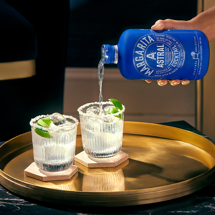 Serving Astral Margarita cocktail from a blue bottle into two glasses with lime garnish.
