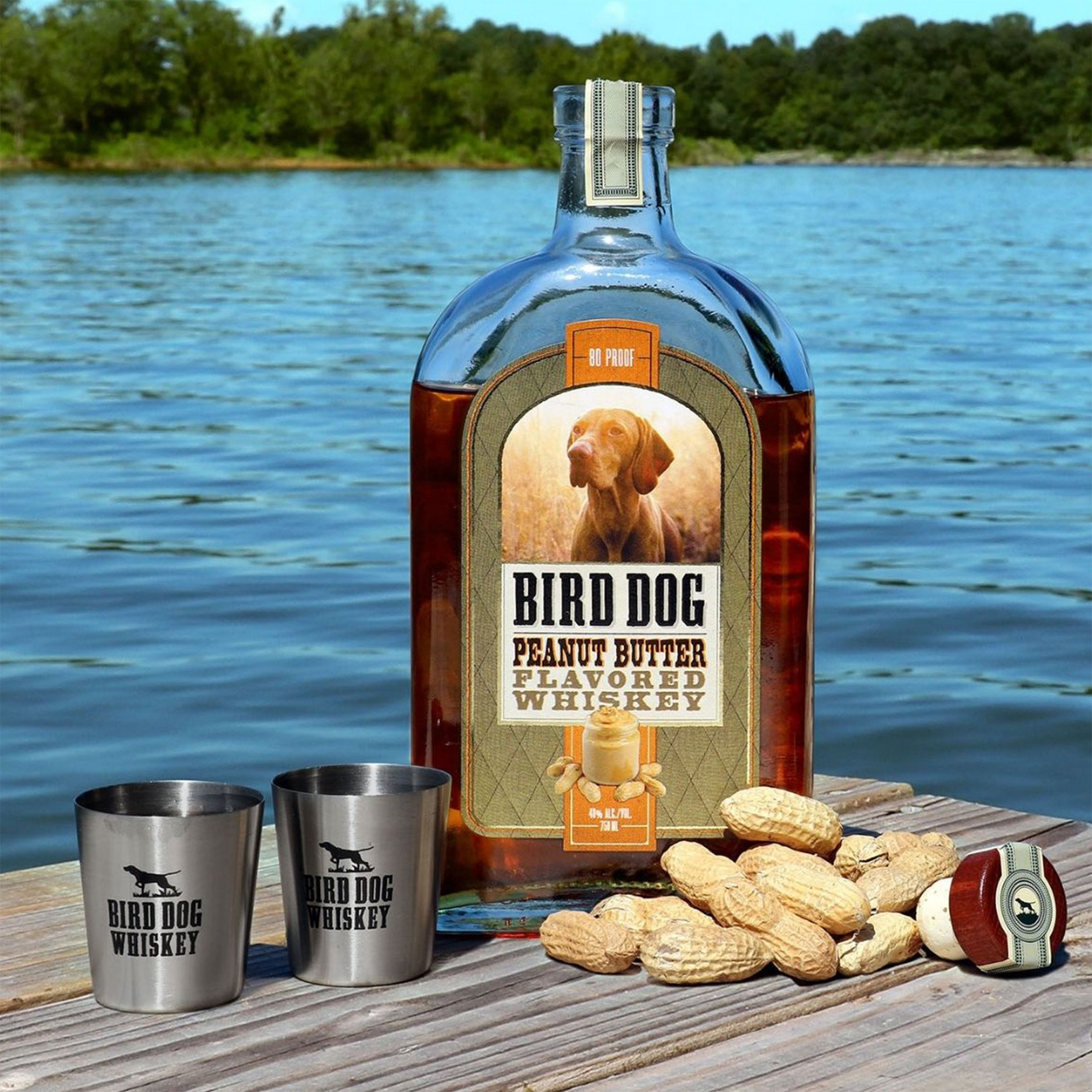 Bird Dog Peanut Butter Flavored Whiskey bottle with shot glasses and peanuts by a lakeside.