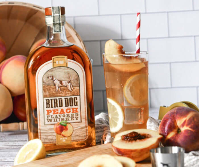 Bird Dog Peach Whiskey bottle with a peach cocktail, garnished with lemon and fresh peaches on a wooden table.