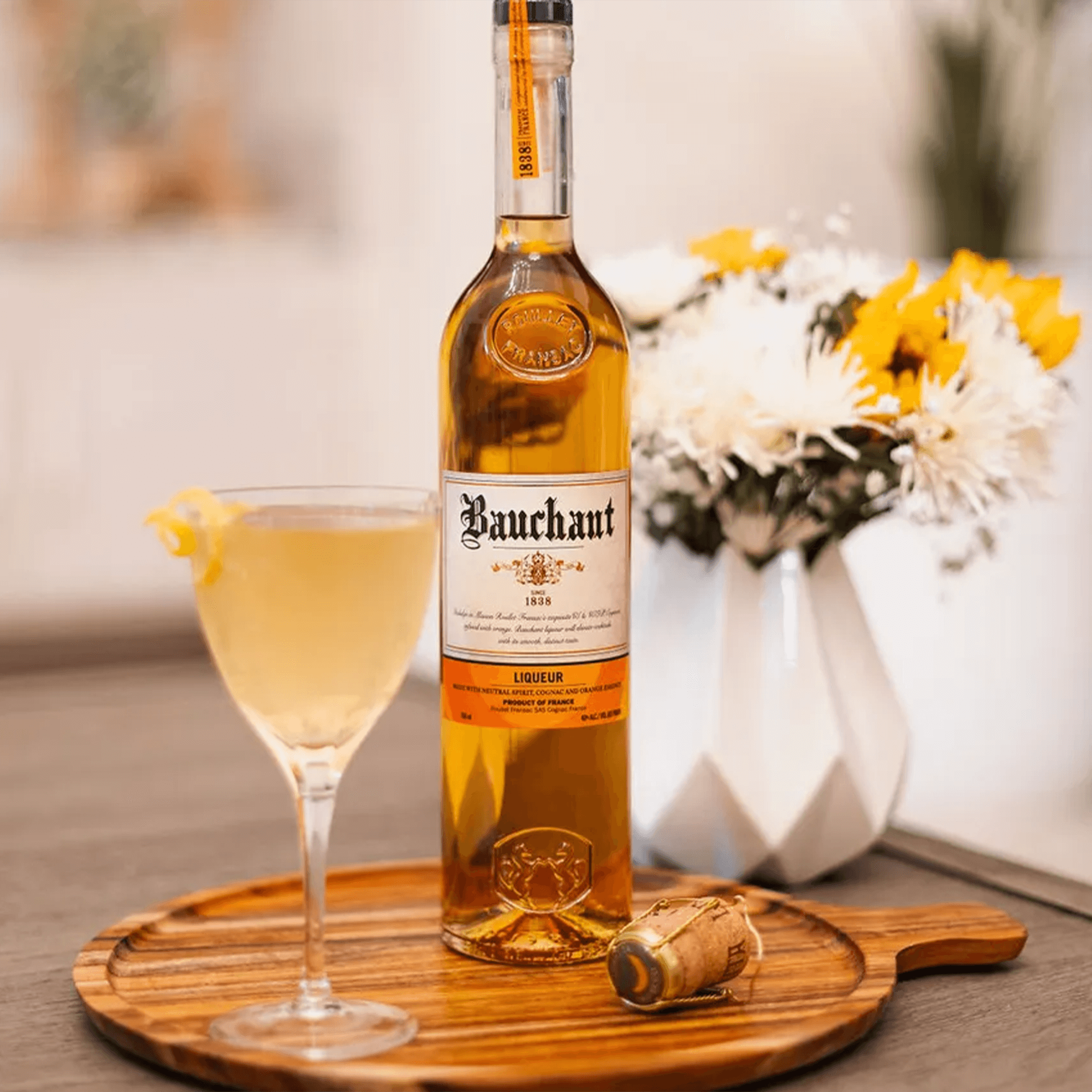 Bauchant Orange Liqueur bottle with cocktail in glass on wooden tray, surrounded by flowers.