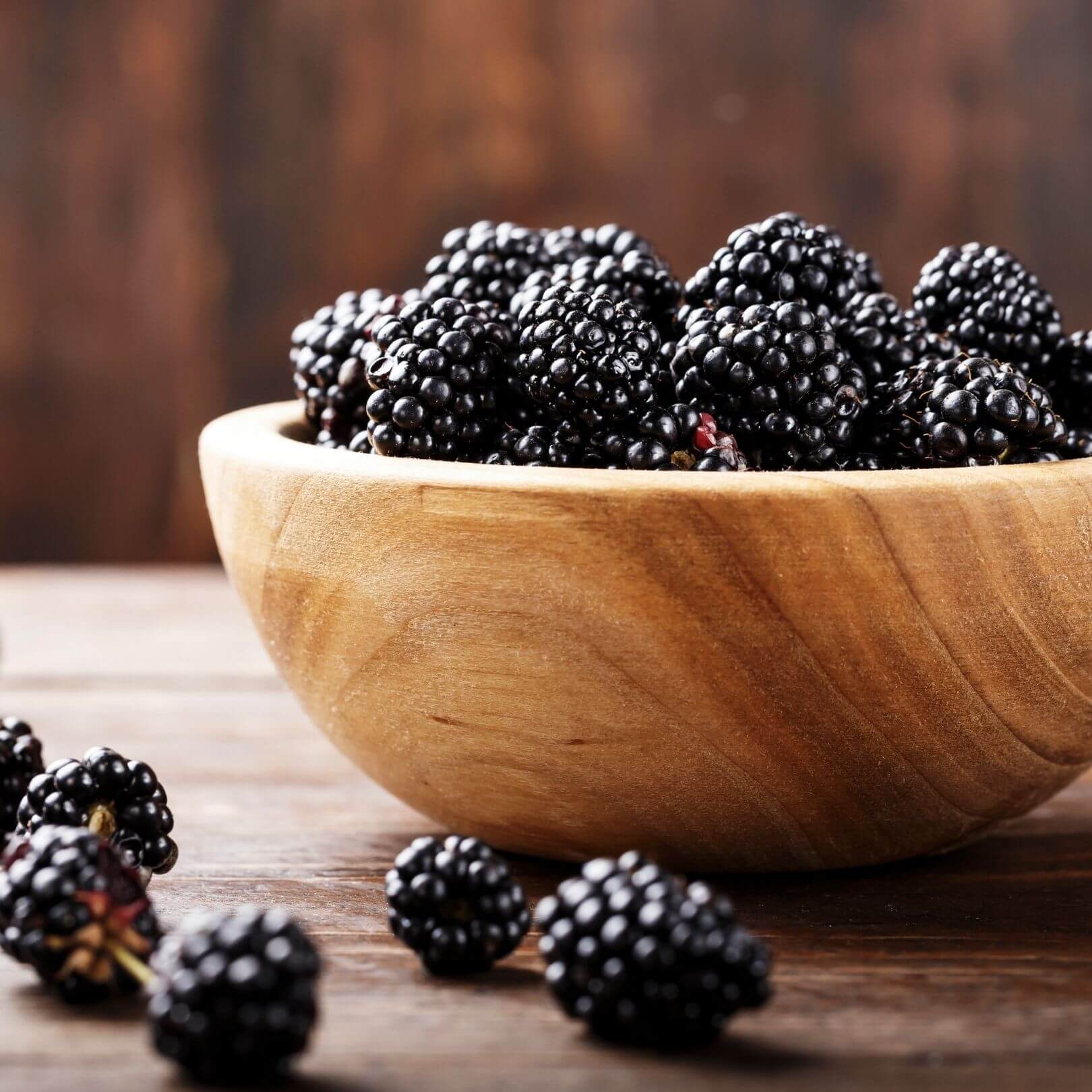 Wooden bowl filled with fresh, ripe blackberries on a rustic wooden surface.