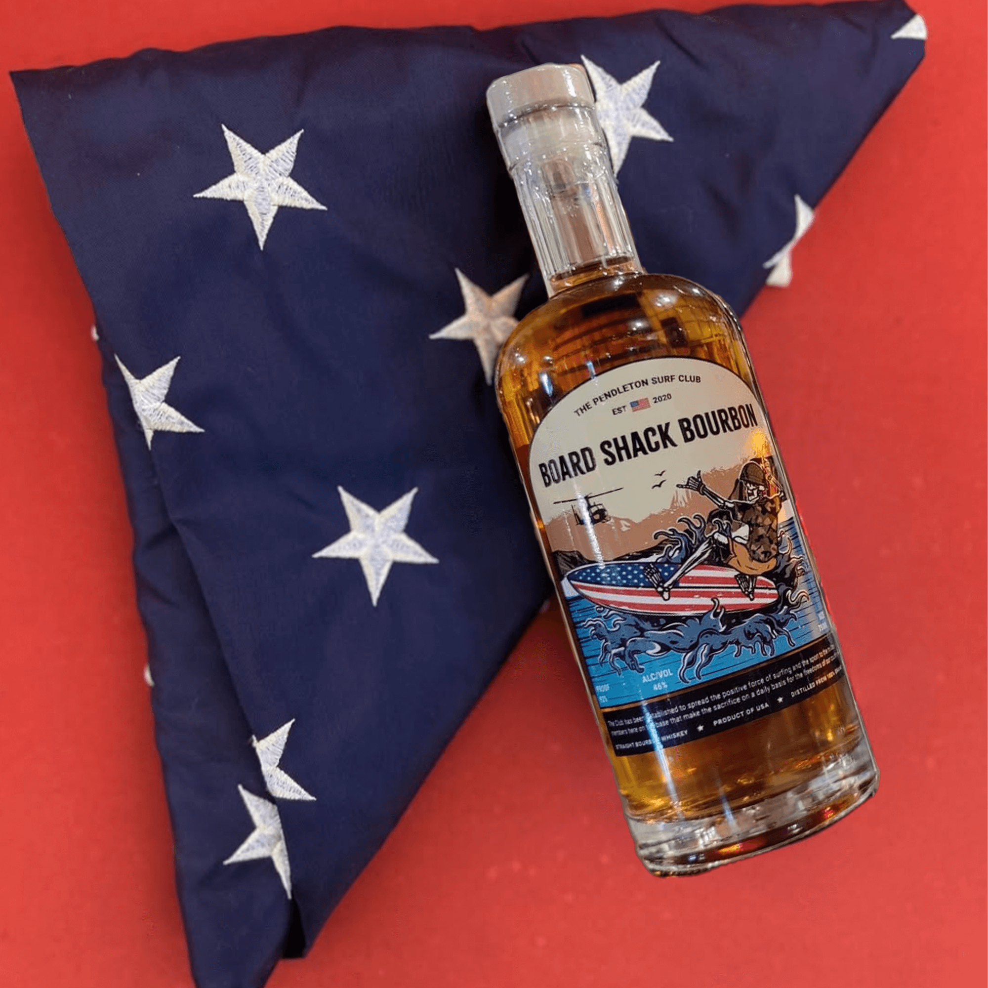Bottle of Board Shack Bourbon displayed with a patriotic star-patterned pillow, representing the Camp Pendleton Surf Club.