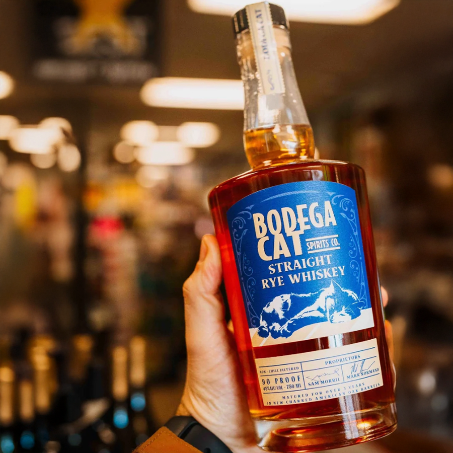 Bodega Cat Straight Rye Whiskey bottle held in hand, showcasing the label and amber color behind a blurred store background.