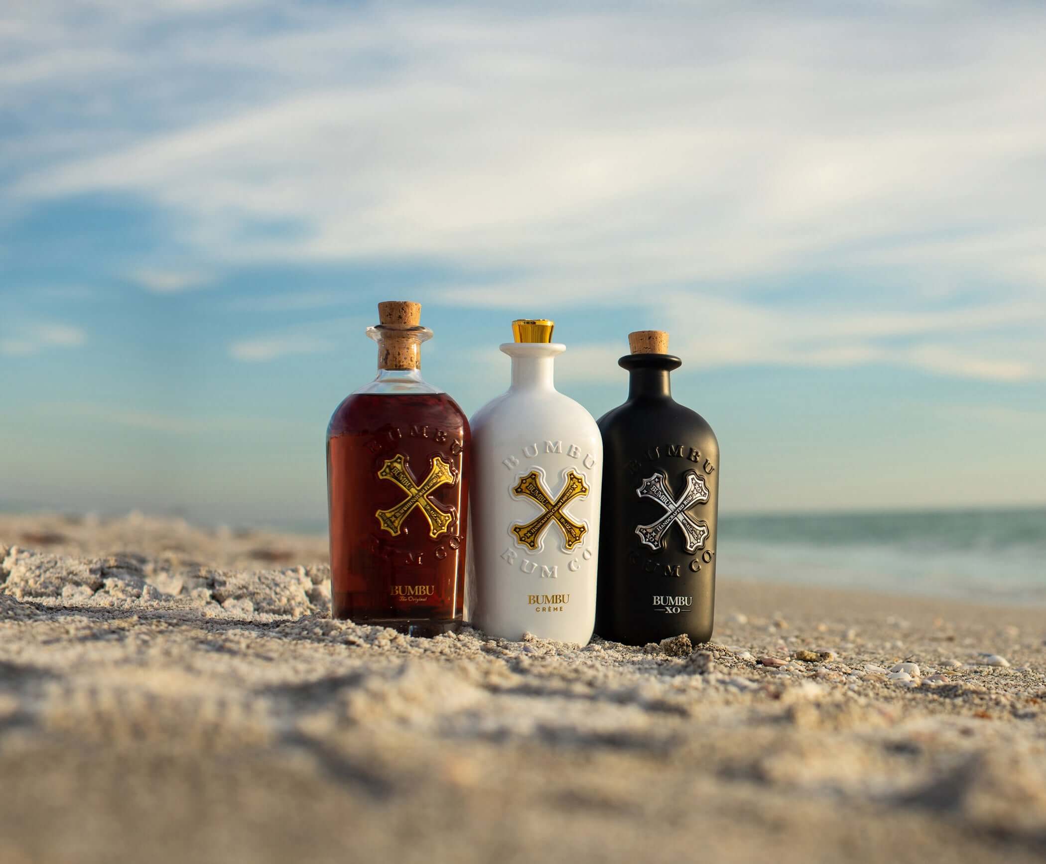 Three Bumbu Rum bottles in various colors on a beach with a blue sky in the background.
