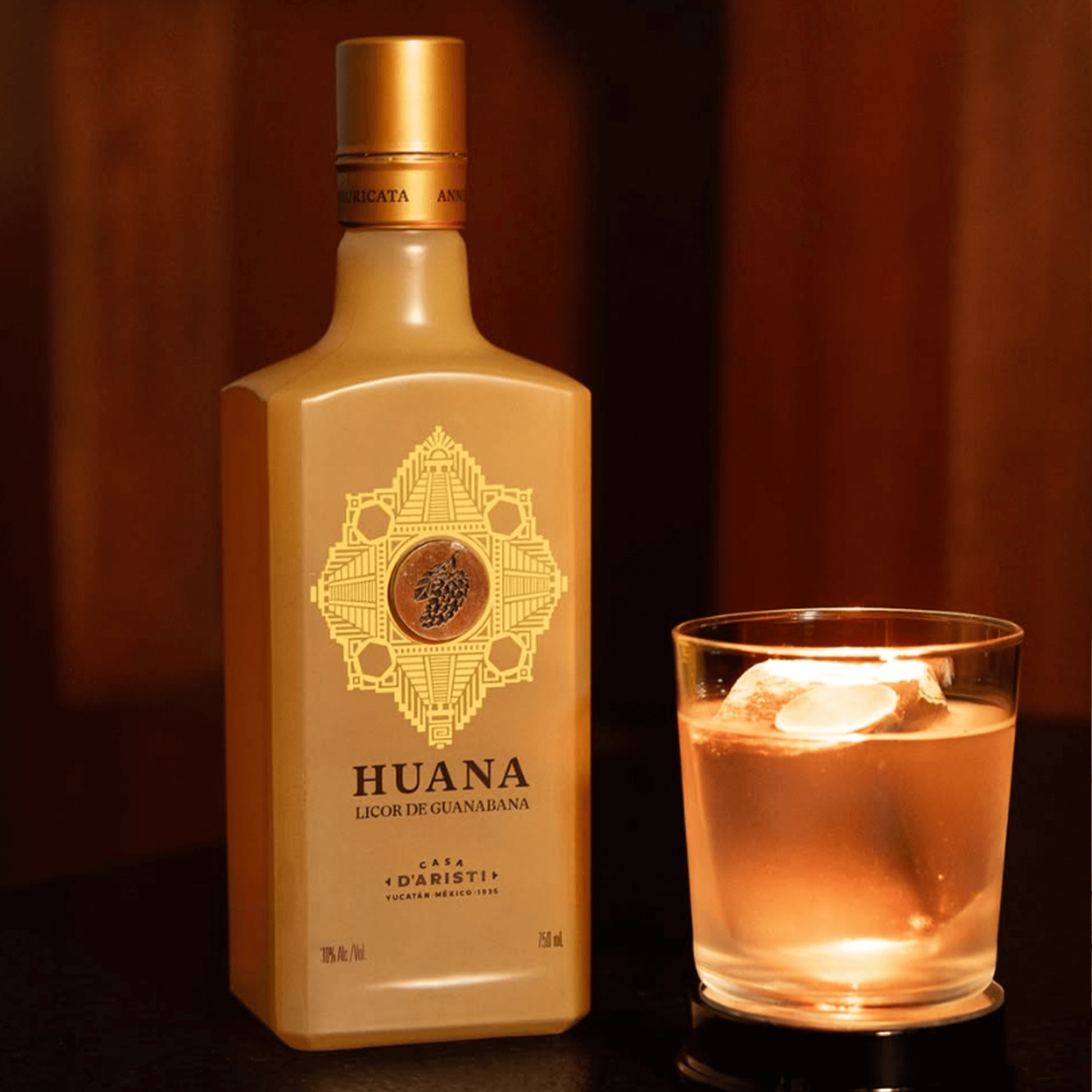 Casa D'Aristi Huana Guanabana liqueur bottle next to a glass of cocktail with ice, set against a dark background.