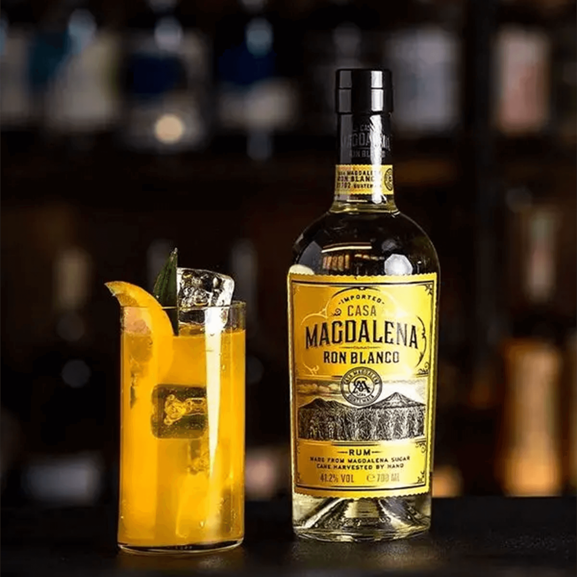 Casa Magdalena Ron Blanco Rum bottle next to a refreshing cocktail with orange and mint on a dark bar.