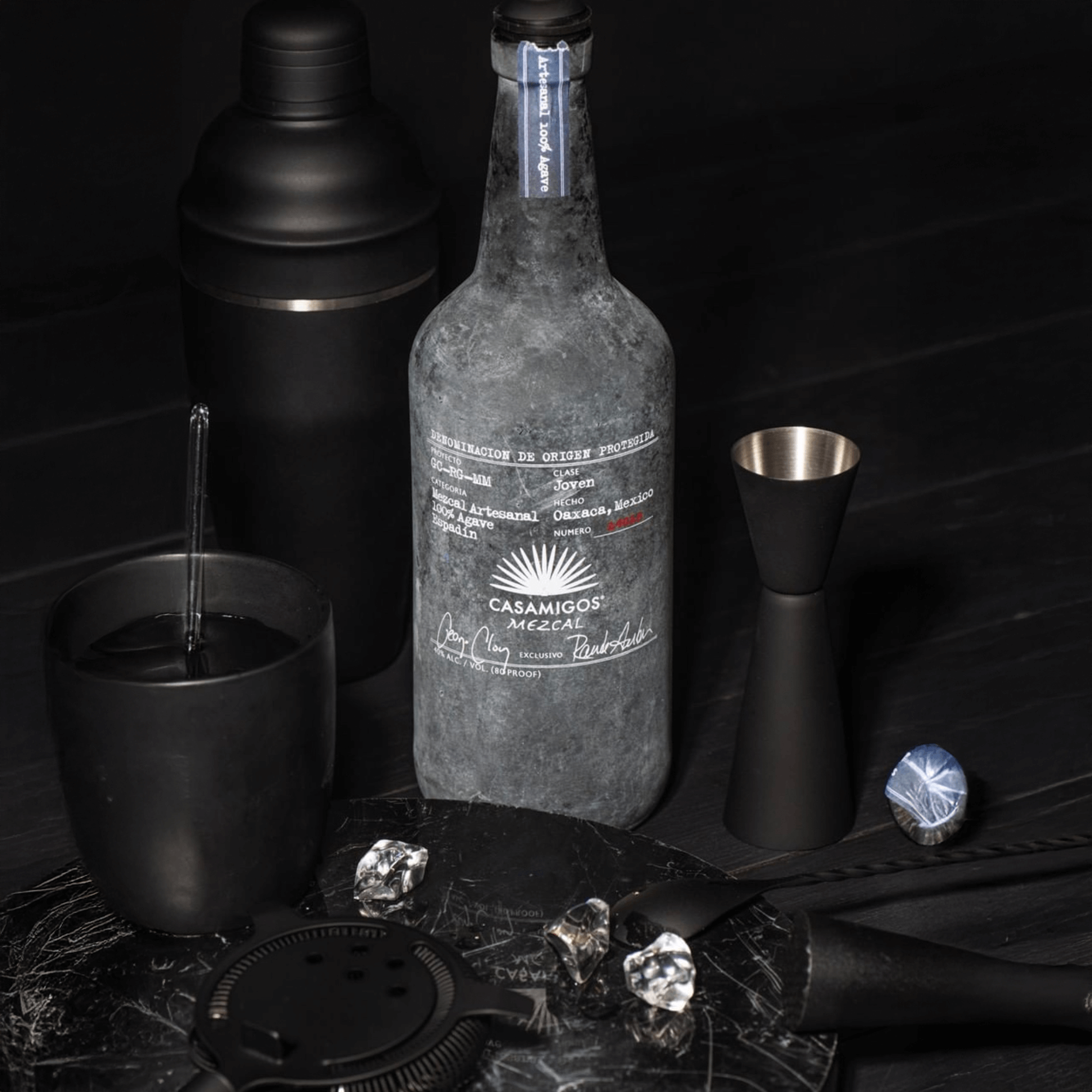 Casamigos Mezcal 1L bottle surrounded by cocktail tools and garnishes on a dark surface.