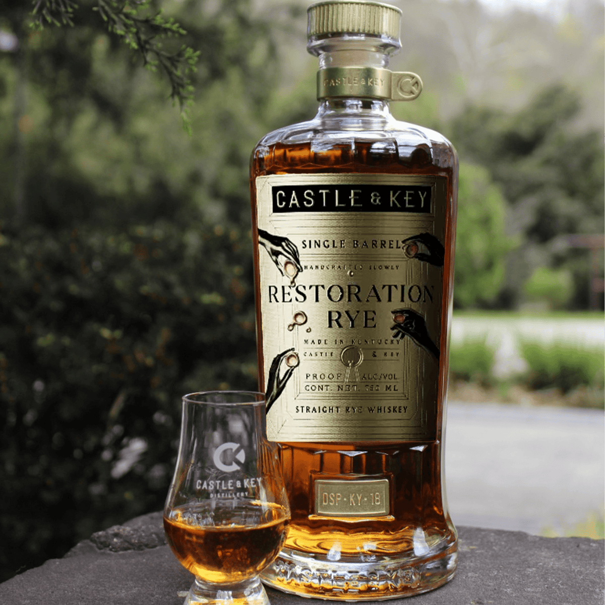 Castle & Key Restoration Single Barrel Rye Whiskey bottle with a tasting glass outdoors, showcasing its rich amber color.