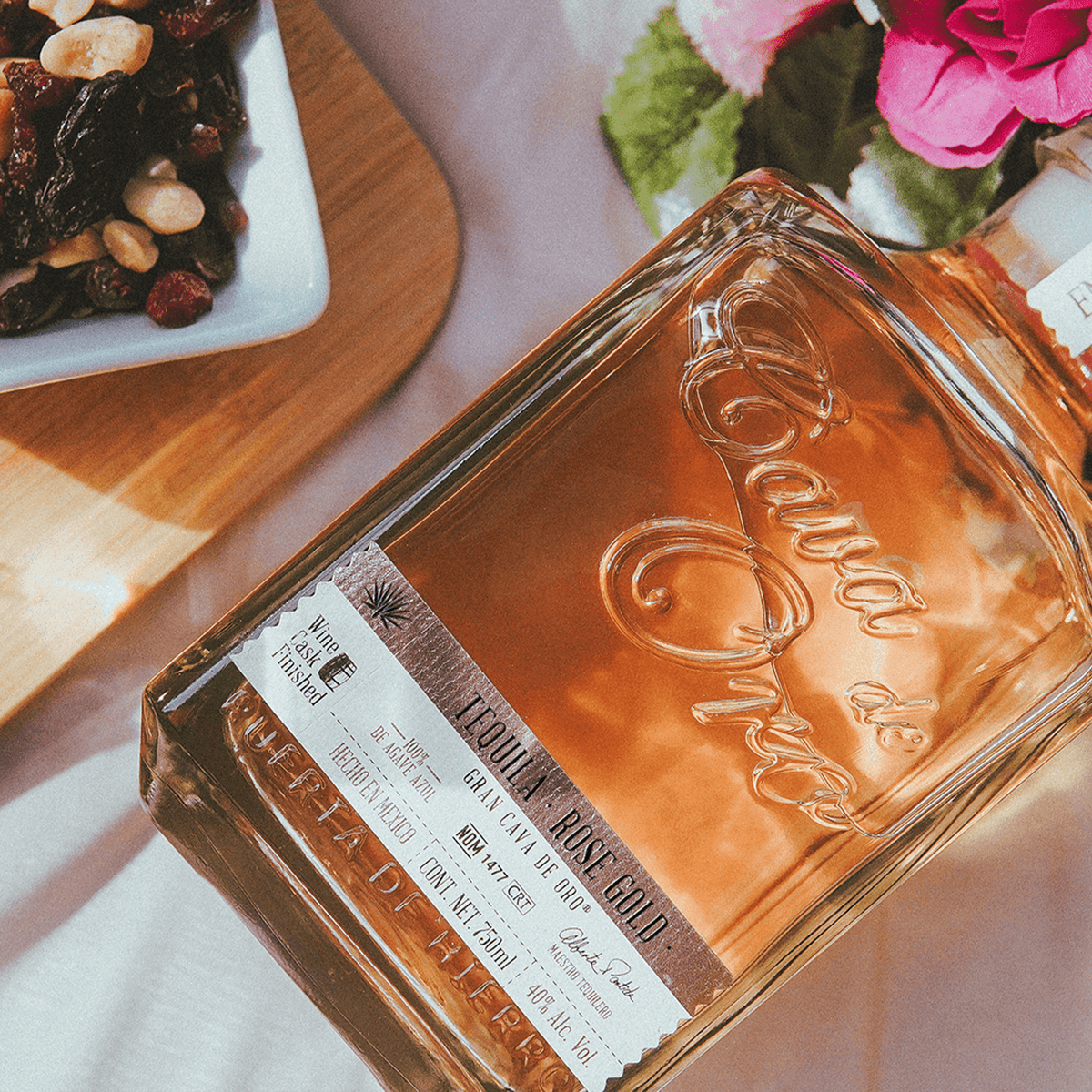 Cava de Oro Rose Gold Tequila bottle with floral backdrop and mixed dried fruits, highlighting its unique flavor profile.