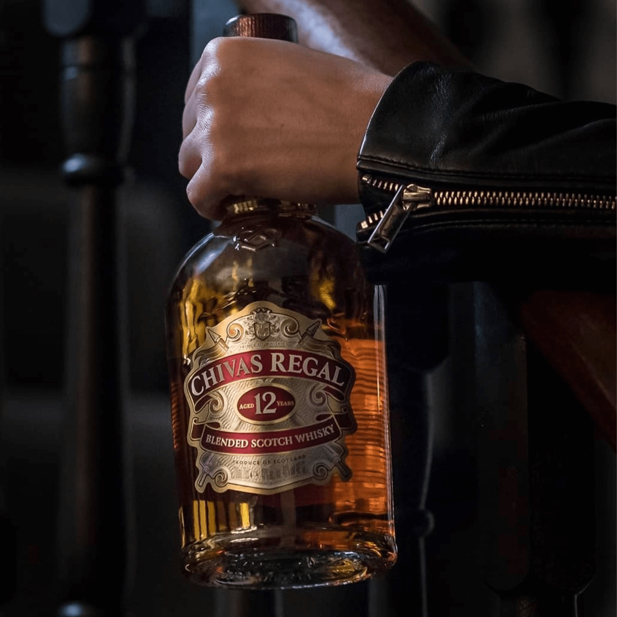 Chivas Regal 12 Year Blended Scotch Whisky bottle held with a leather jacket in a dimly lit setting.