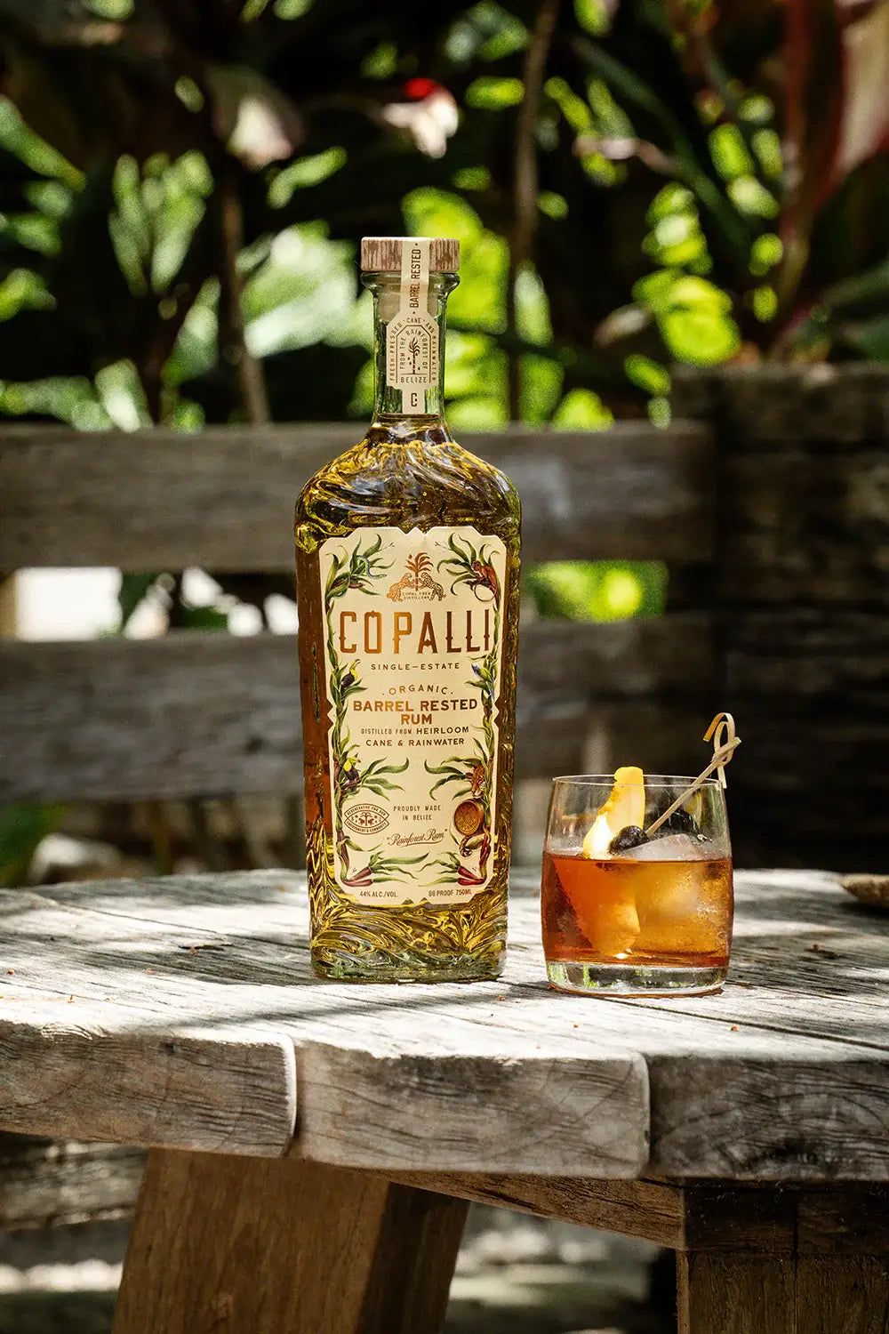 Copalli Barrel Rested Belizean Rum bottle and cocktail on a wooden table in a lush outdoor setting.