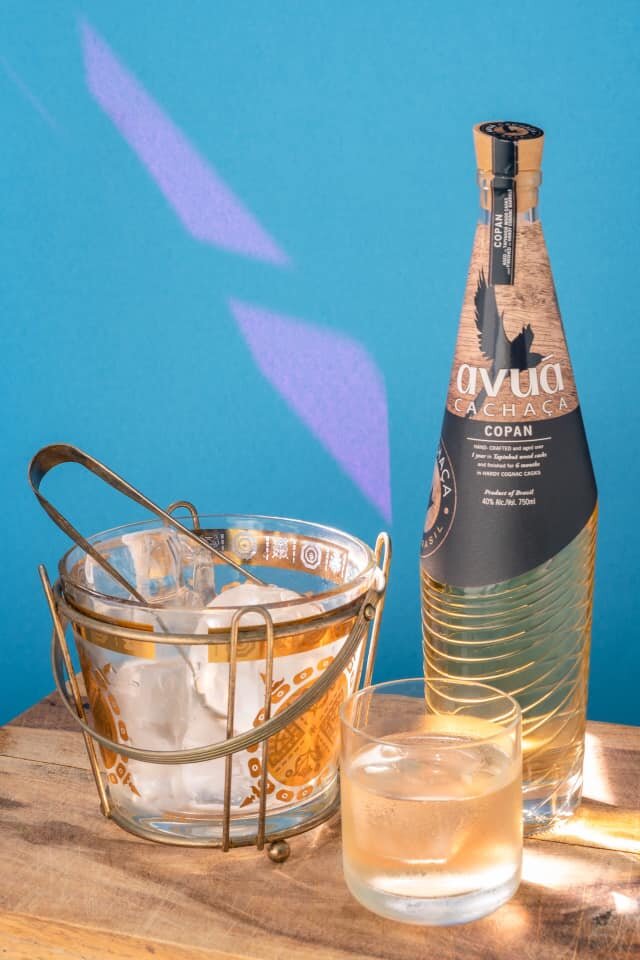 Avuá Copan Limited Edition Aged Cachaça bottle alongside ice bucket and glass, set against a blue background.