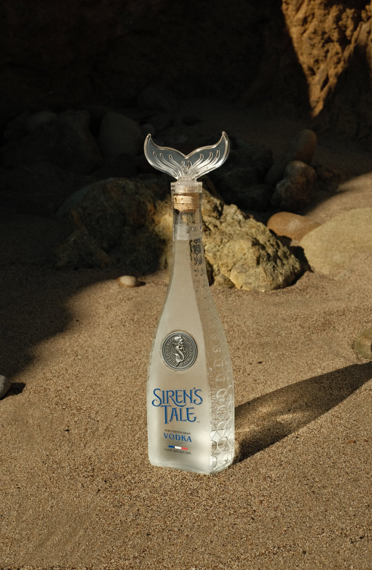Siren's Tale French Vodka limited edition bottle on sandy beach, featuring an elegant design and unique tail-shaped cap.