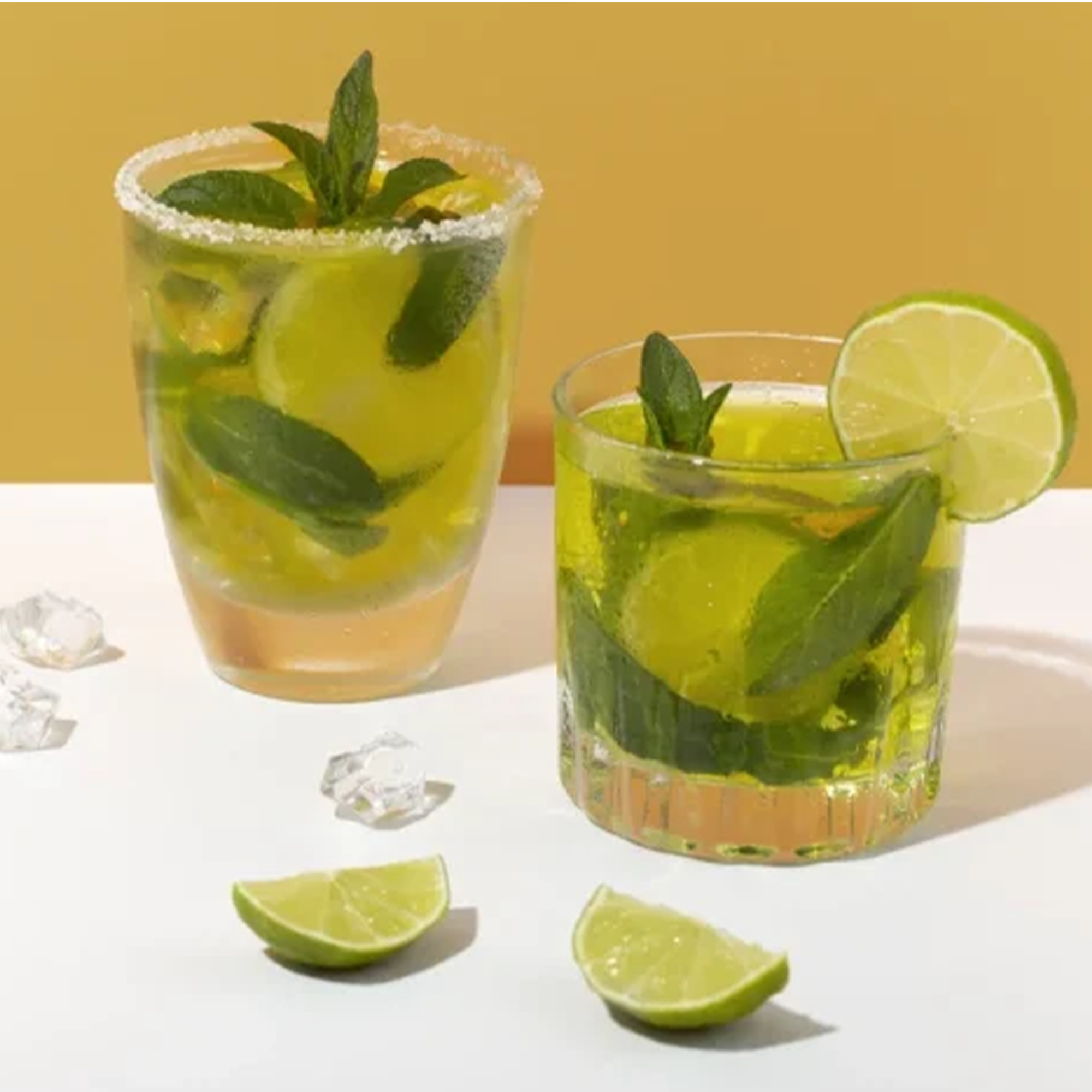 Two refreshing cocktails with mint, lime slices, and ice against a vibrant yellow background.