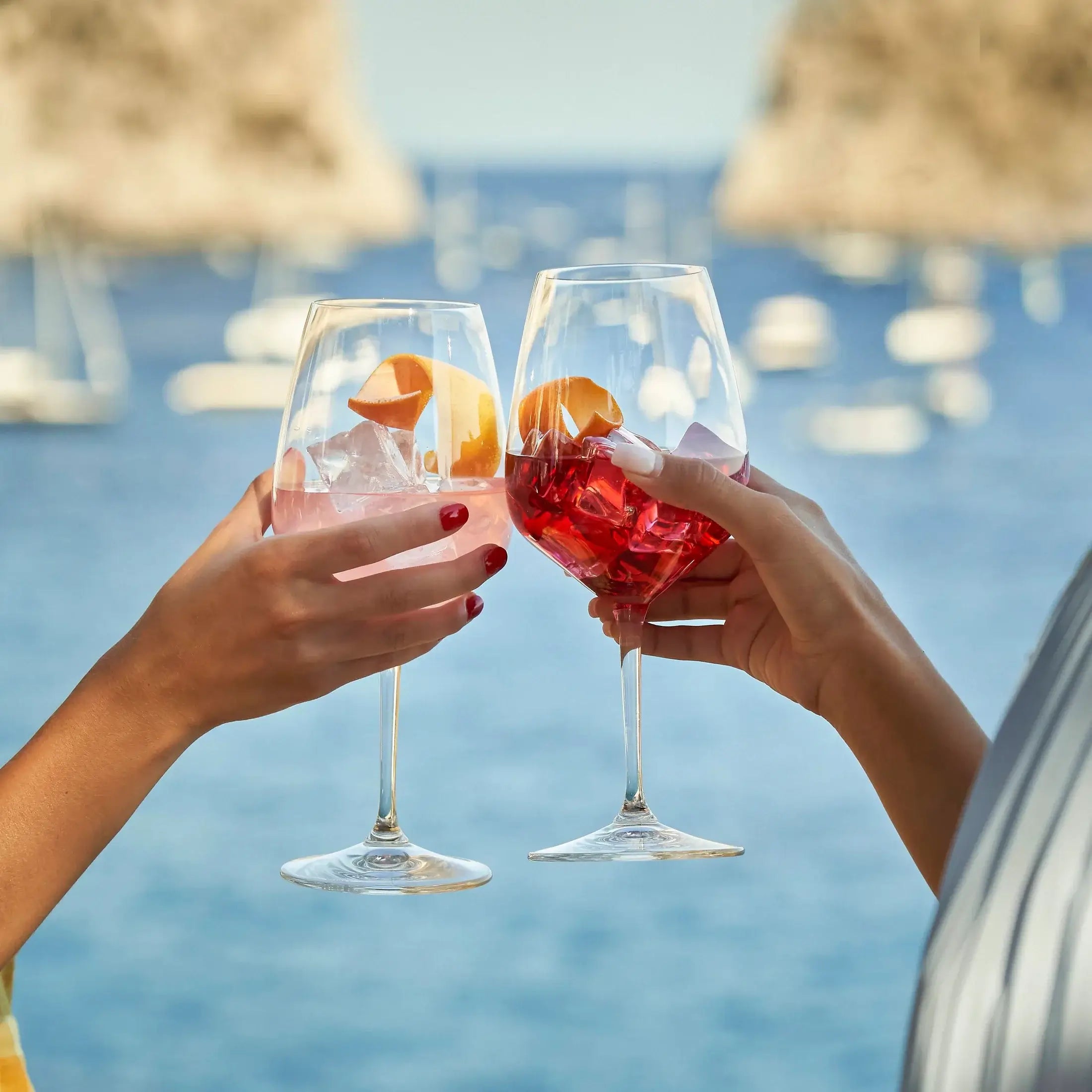 Two hands clinking glasses of Delola Paloma Rosa Spritz against a scenic seaside backdrop.