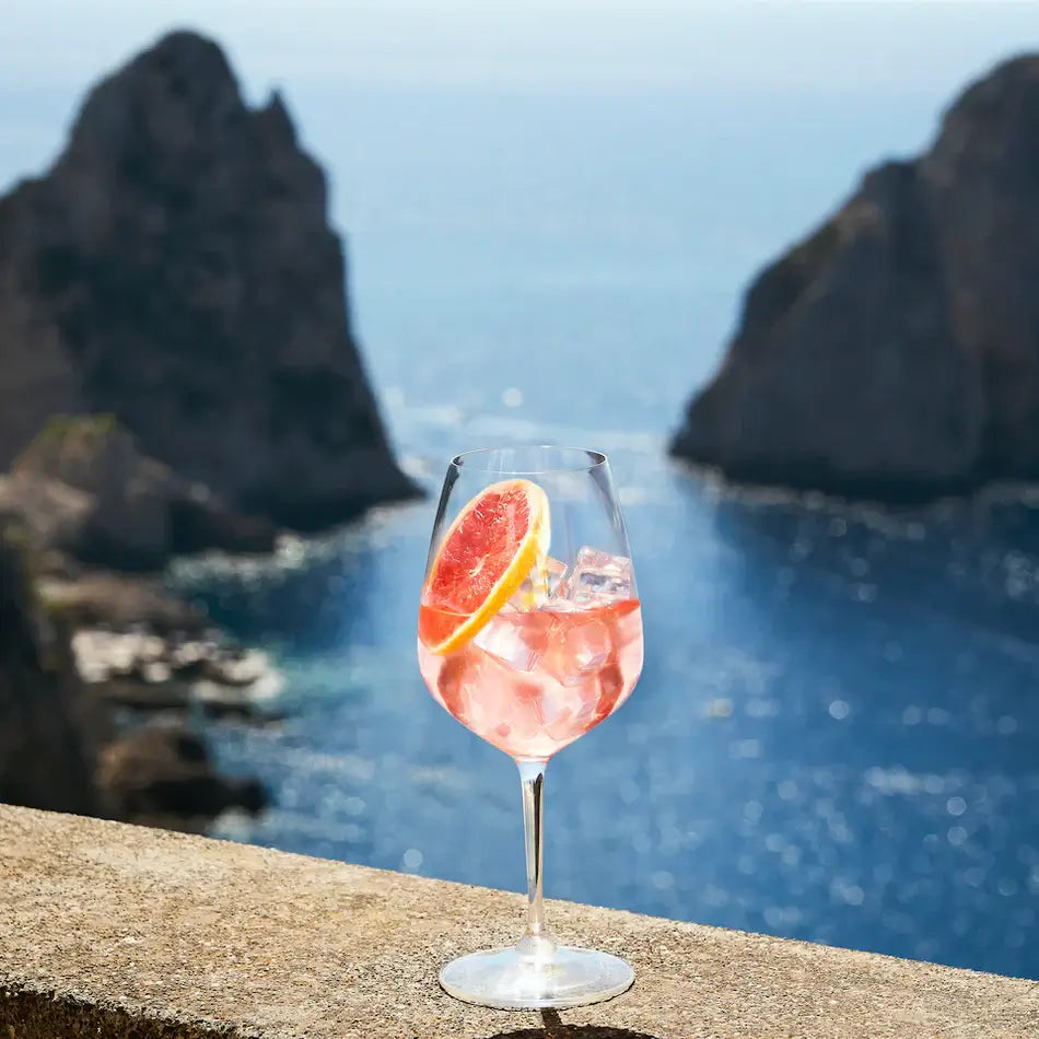 Glass of Delola Paloma Rosa Spritz with a slice of grapefruit overlooking a beautiful blue ocean.