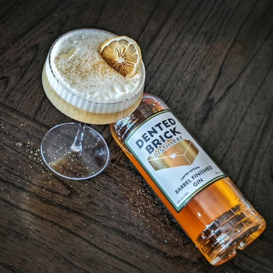 Dented Brick Barrel Finished Gin bottle and cocktail on wooden table with lemon garnish and spices.