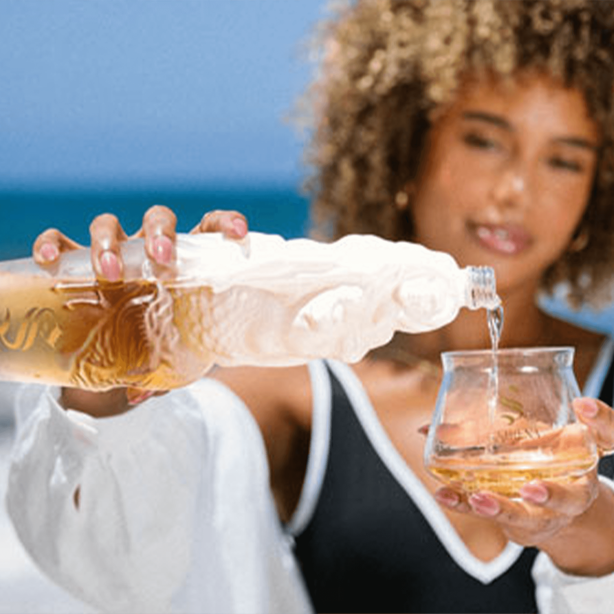 Woman pouring Dia De La Sirena Reposado Tequila from a unique bottle into a glass by the beach.