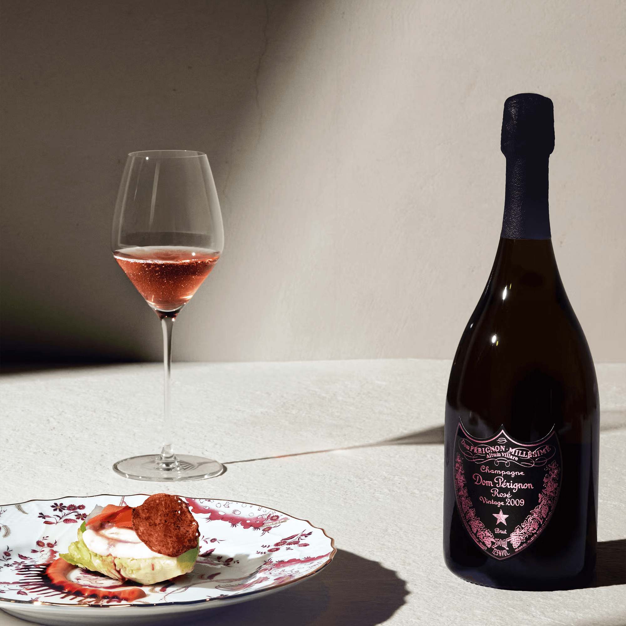 Dom Pérignon Rosé 2009 bottle next to a glass of rosé wine and a gourmet dish on a decorative plate.