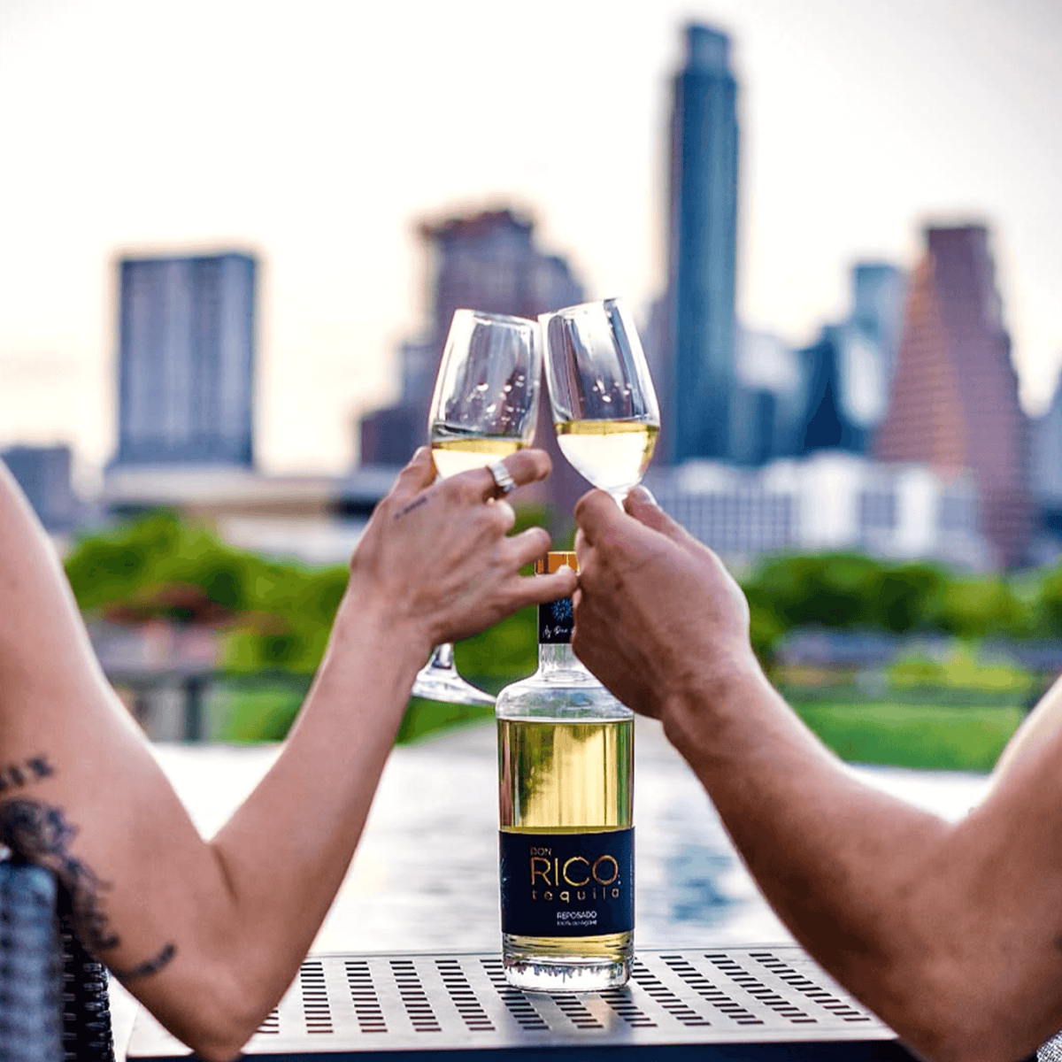 Glasses clinking over Don Rico Reposado Tequila bottle with city skyline backdrop, celebrating craftsmanship and tradition.