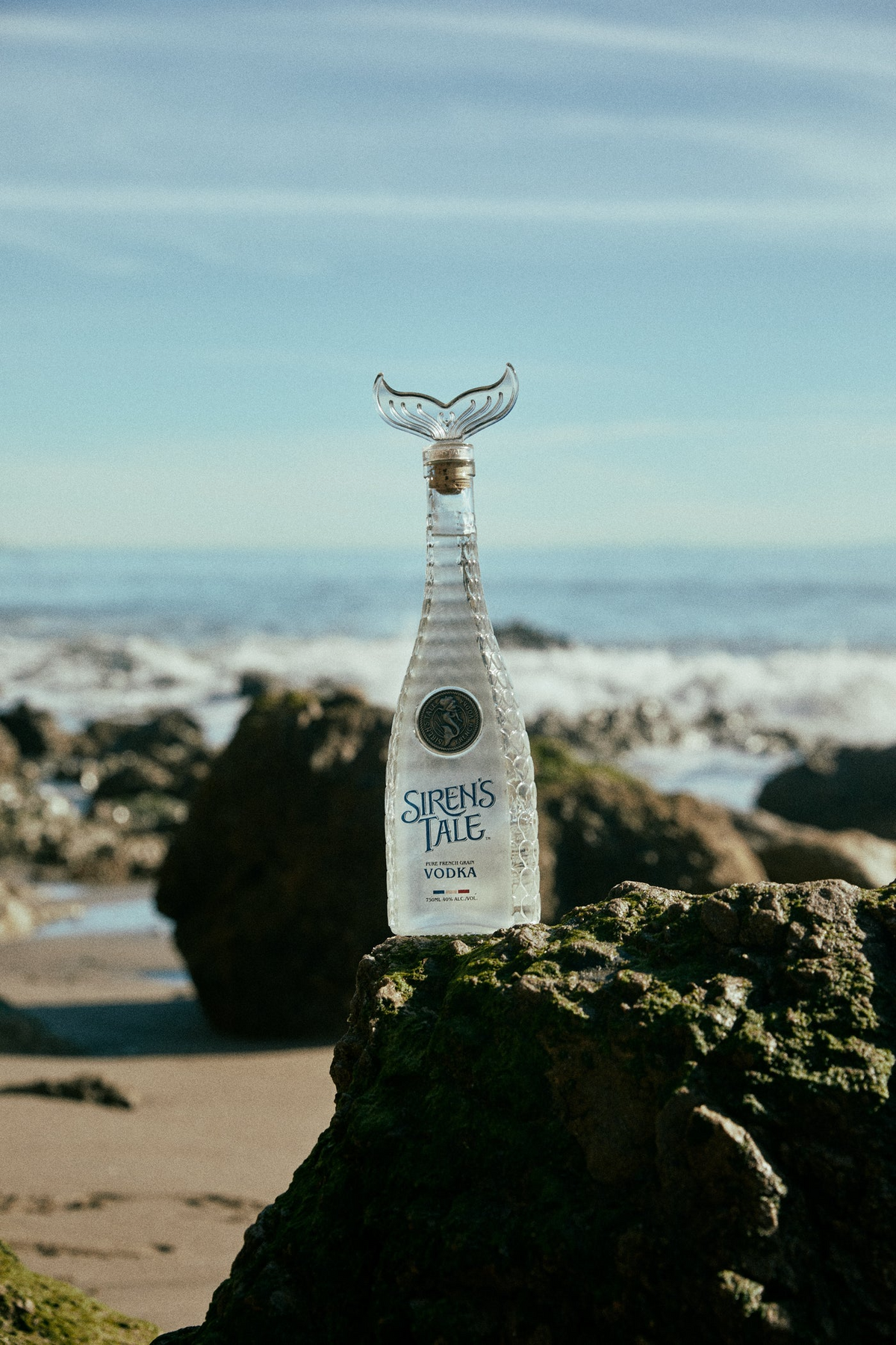 Siren's Tale French Vodka bottle with a whale tail cap on a rocky beach.