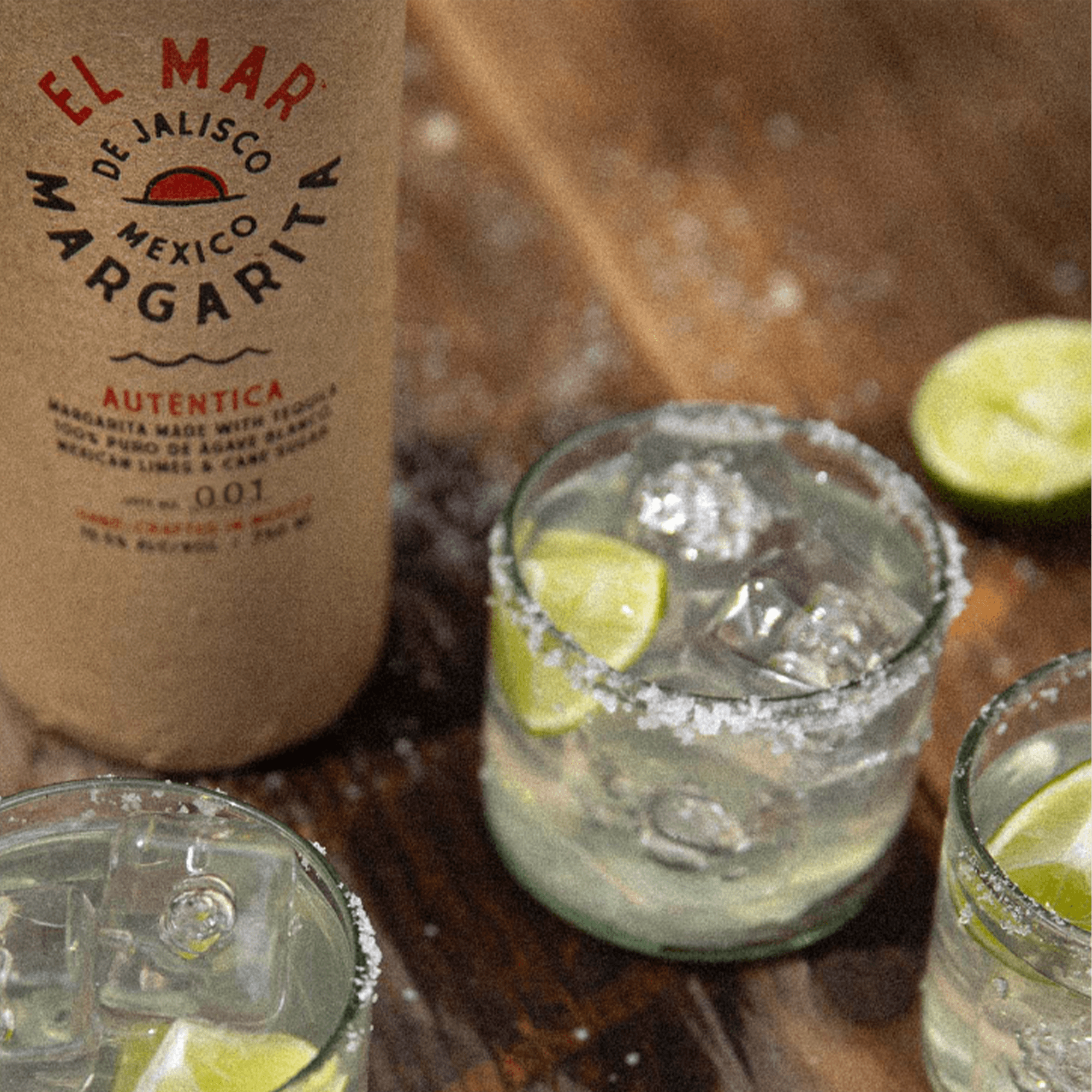 El Mar Margarita bottle beside glasses with margarita, ice, lime slices on wooden table.