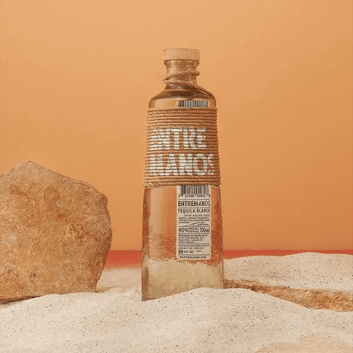 Entremanos Blanco Tequila 700ml bottle set against a warm orange background with sandy stones.