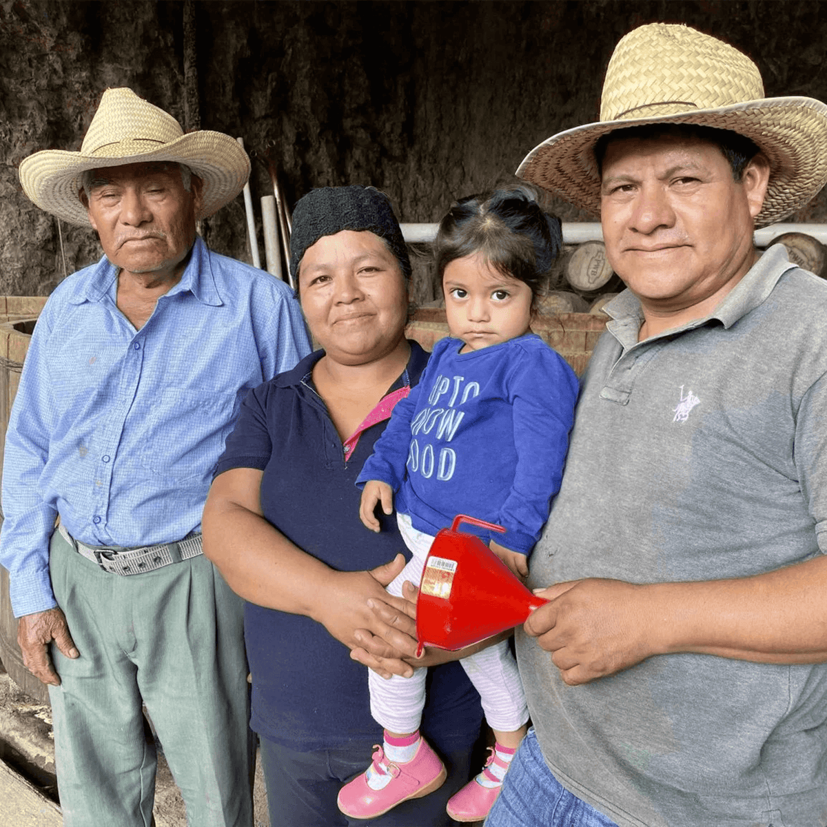 Four individuals in traditional attire with straw hats, standing together in a rustic setting, holding a small child.