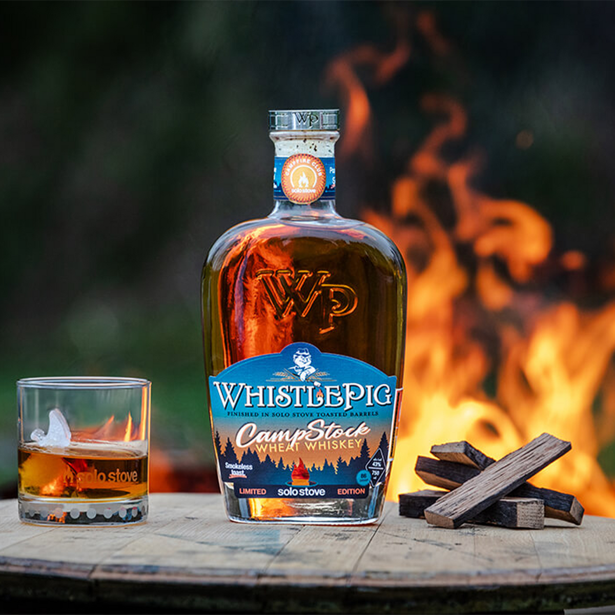 WhistlePig CampStock Wheat Whiskey bottle with glass beside a campfire, showcasing summer vibes and flavor notes.