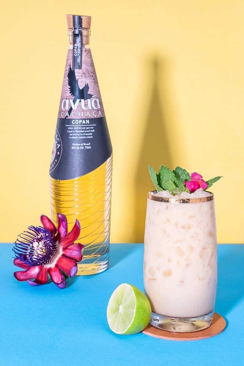 Avuá Copan Limited Edition Aged Cachaça bottle next to a mixed drink with flowers and lime.