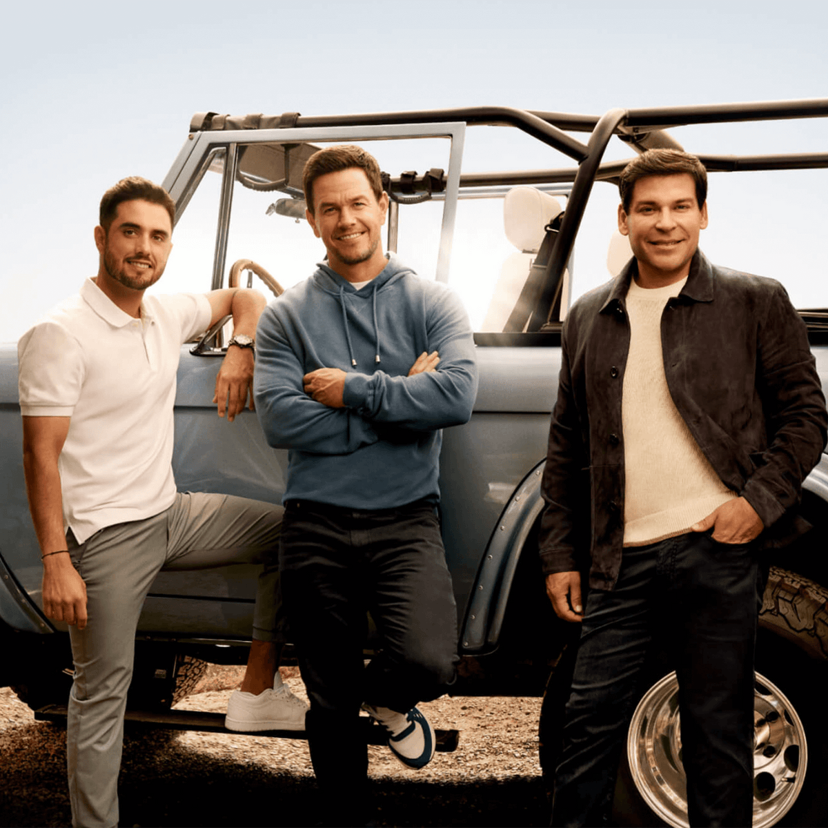 Three men posing by a vintage jeep, featuring a relaxed outdoor vibe and casual attire in a sunny setting.
