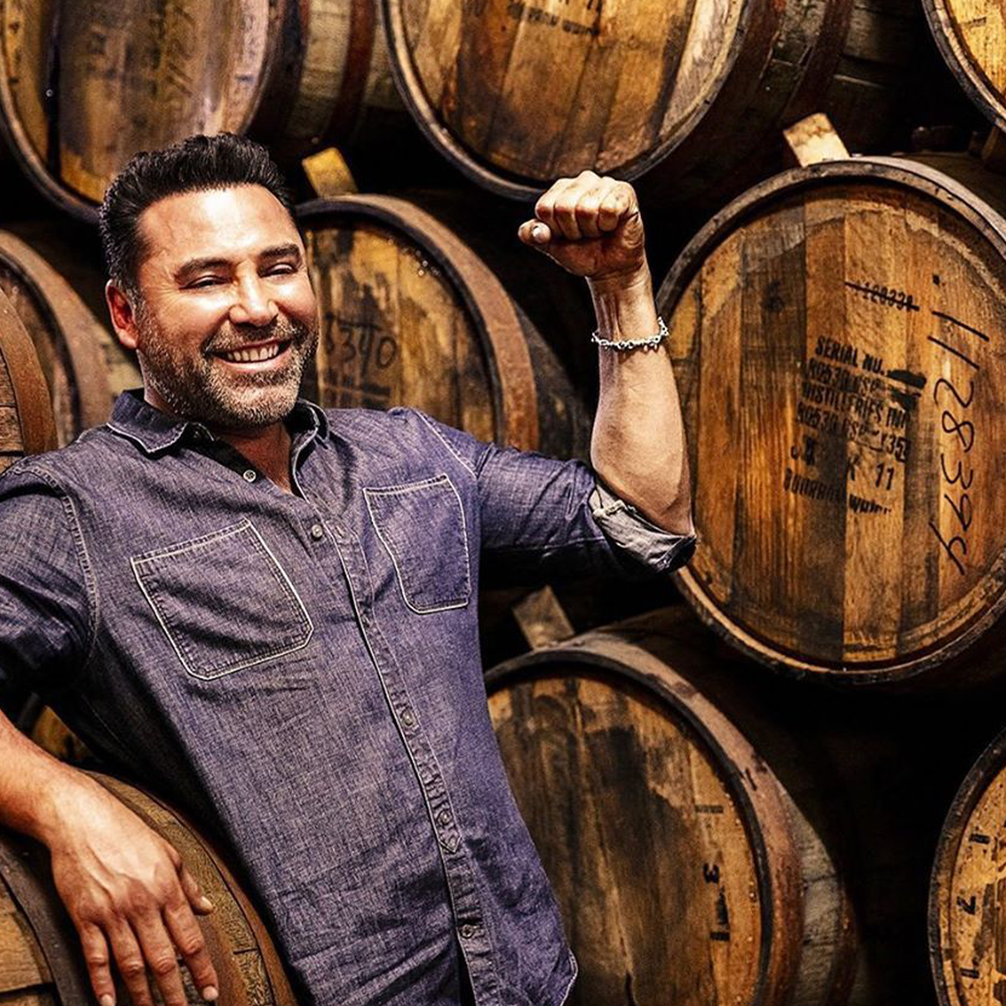 Smiling man in a denim shirt poses beside tequila barrels, showcasing the craftsmanship behind Casa Mexico Blanco Tequila.