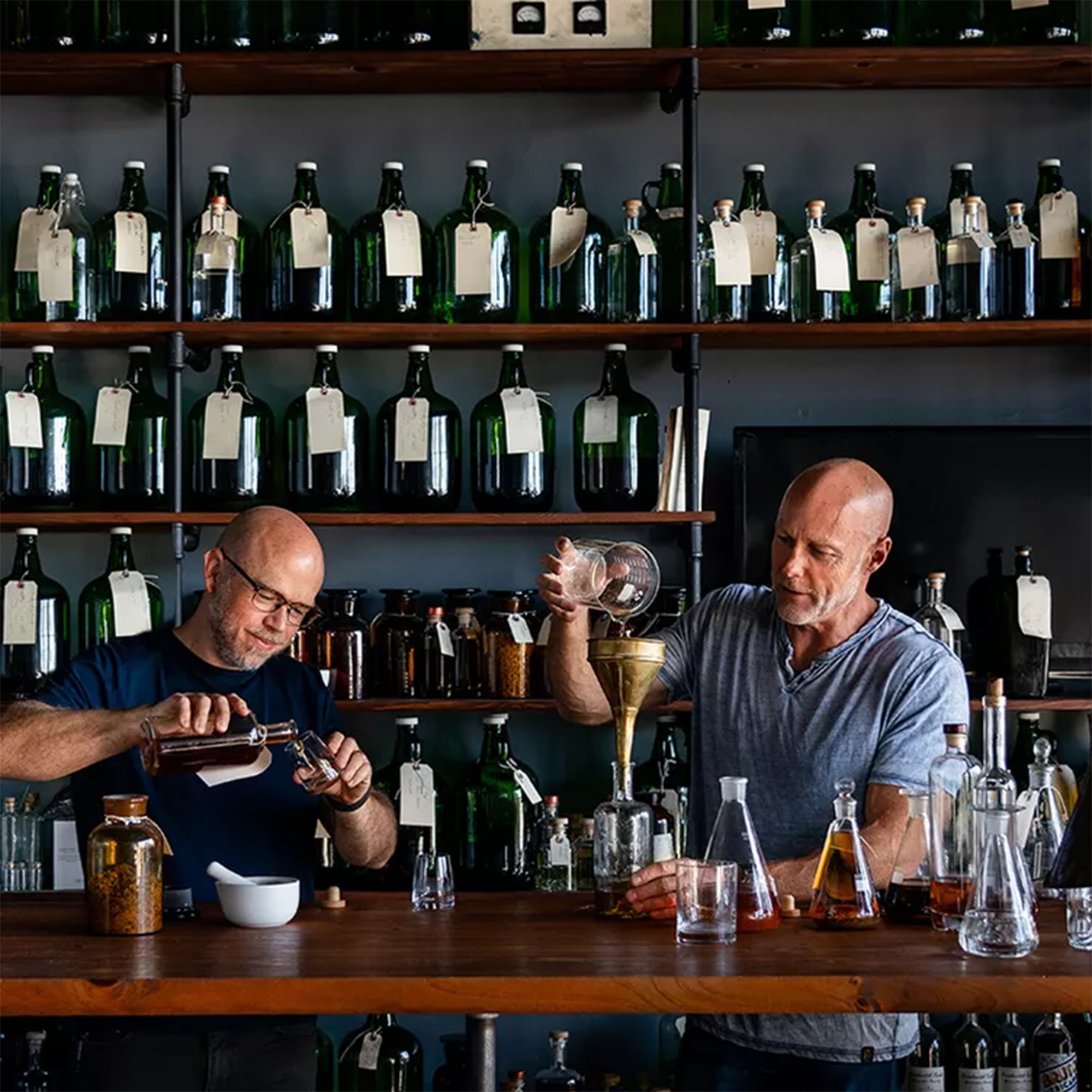 Bartenders expertly mixing and pouring spirits at a bar with shelves of green bottles in the background.